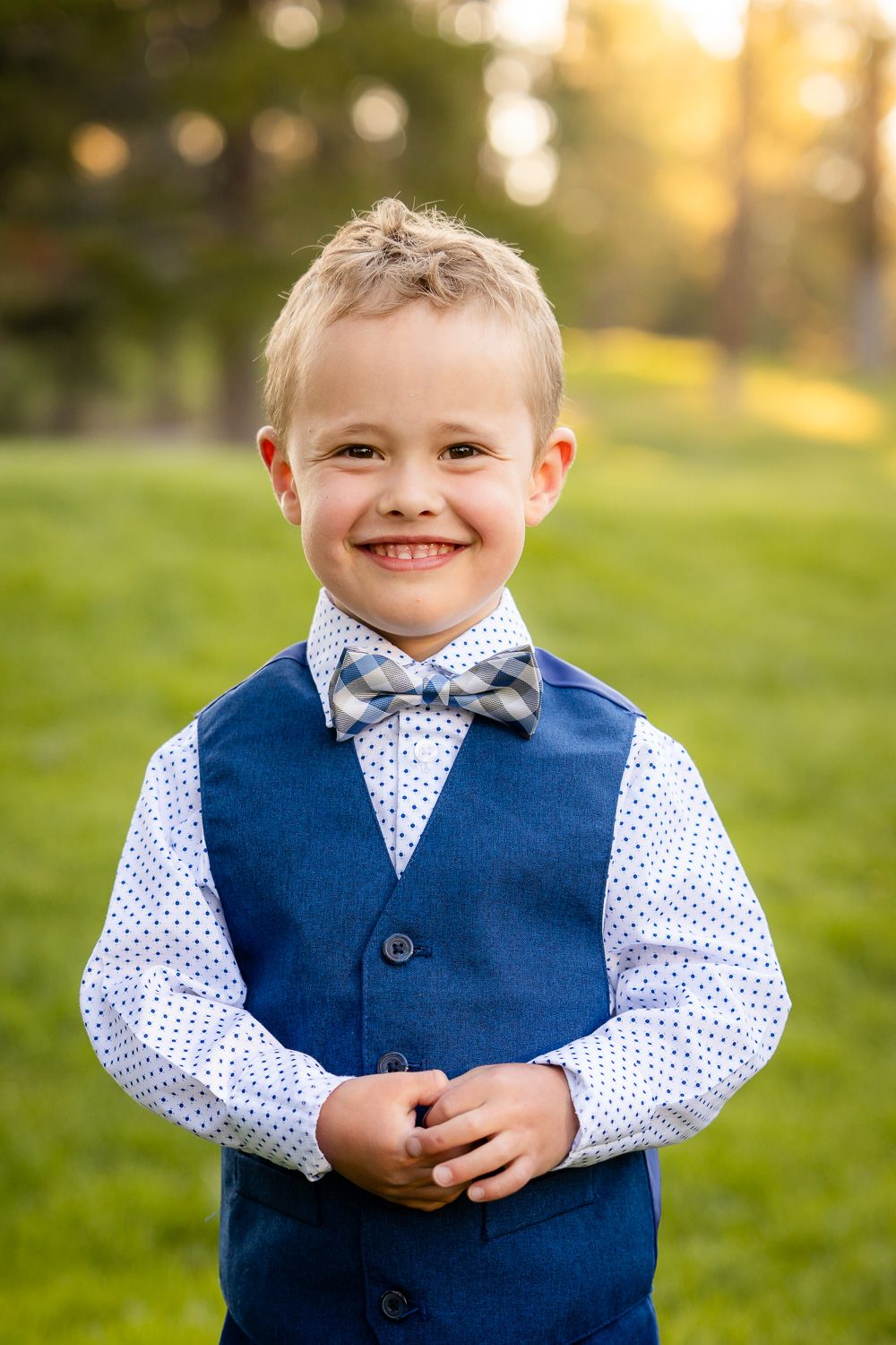 A dressed-up young child wearing a blue vest, polka dot shirt and bow tie smiles in an outdoor setting at sunset.