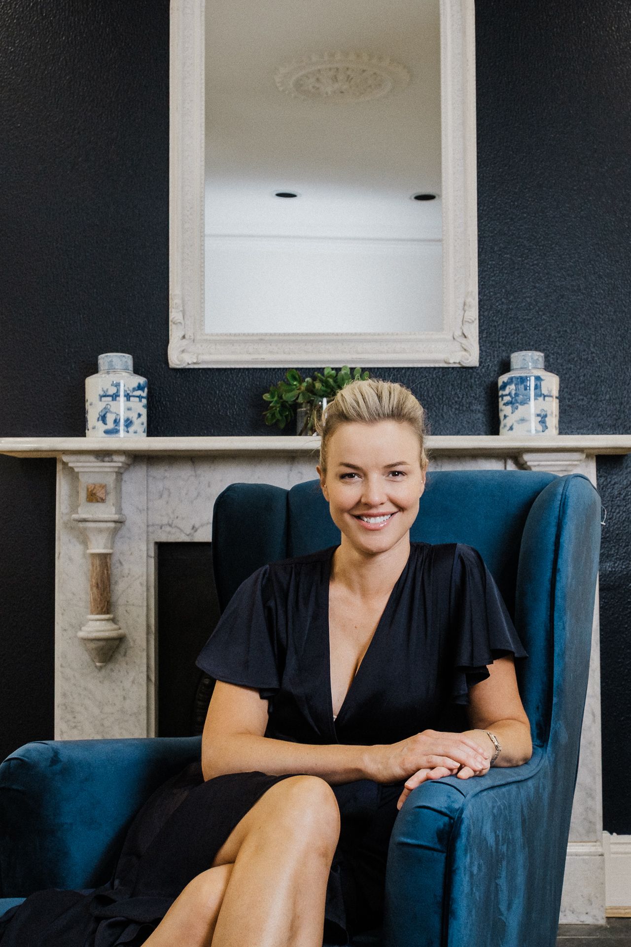 A person in a black dress sits elegantly in a navy blue armchair against a dark wall with decorative mantle.