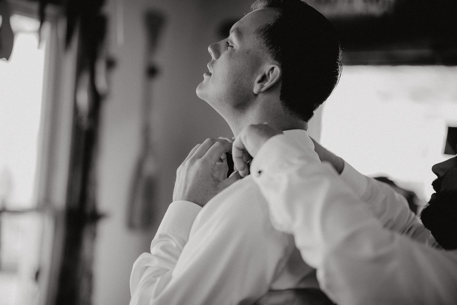 Black and white portrait of person in white shirt adjusting collar while looking up in contemplative pose.