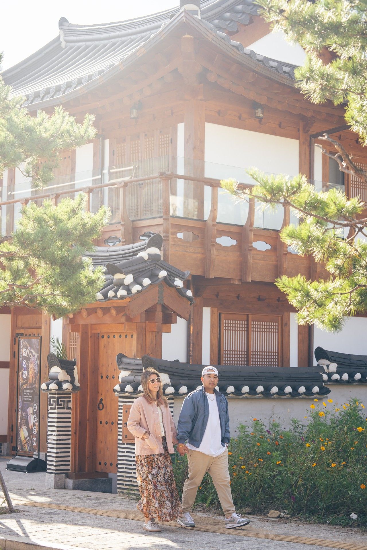 A couple walks past a traditional Korean hanok building with wooden architecture and curved tile roof on a sunny day.