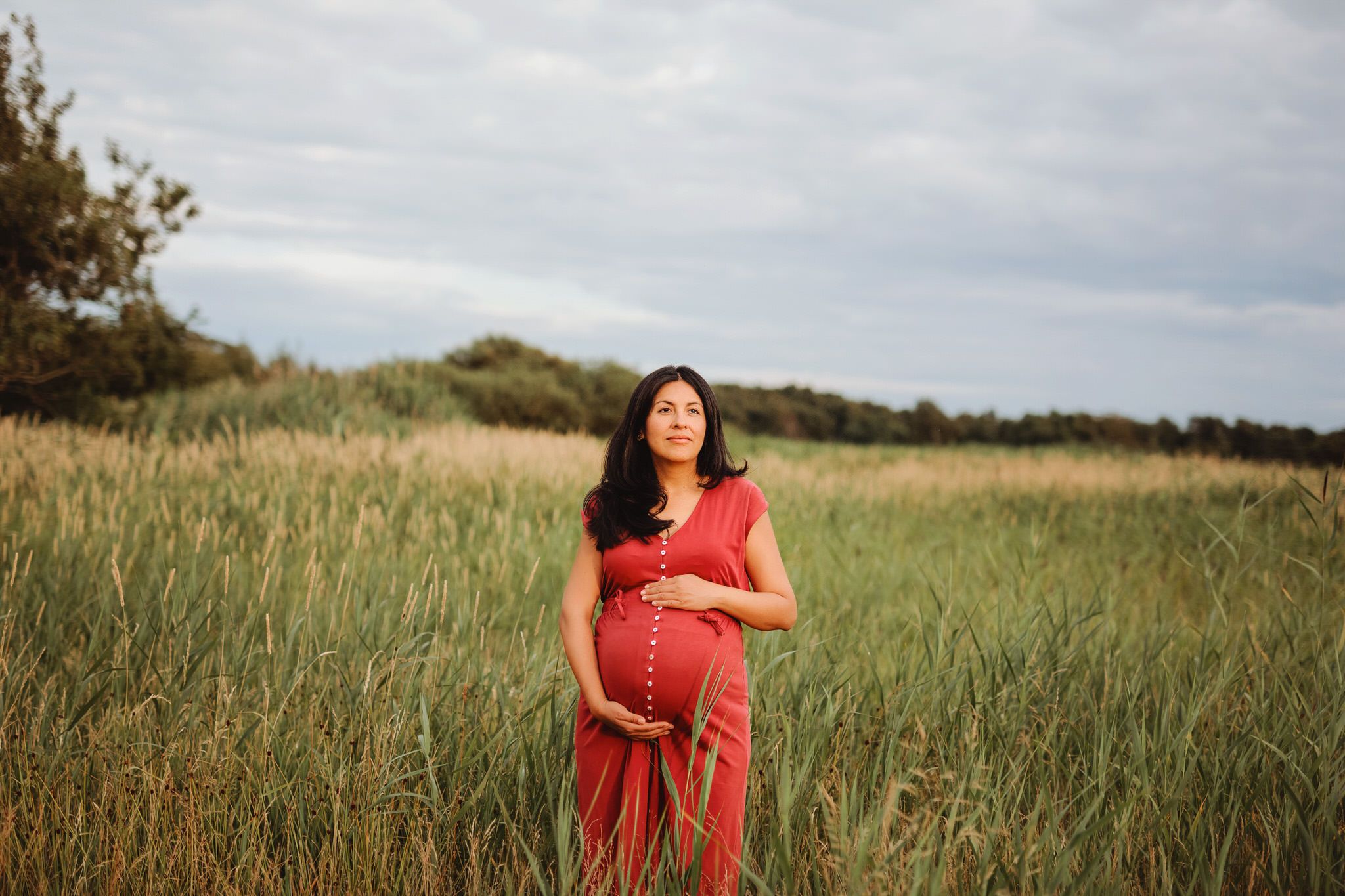 Gravid kvinna håller om sin mage och står i hög gräs. Gravidfotografering.