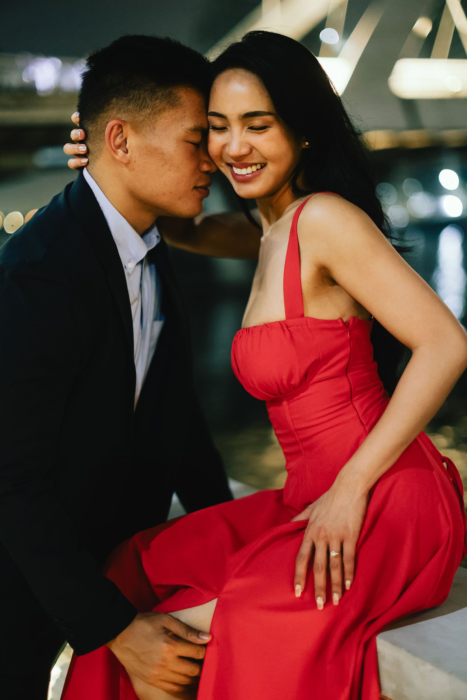 Romantic couple shares intimate moments in an urban setting as she wears a vibrant red dress against dark evening lighting.