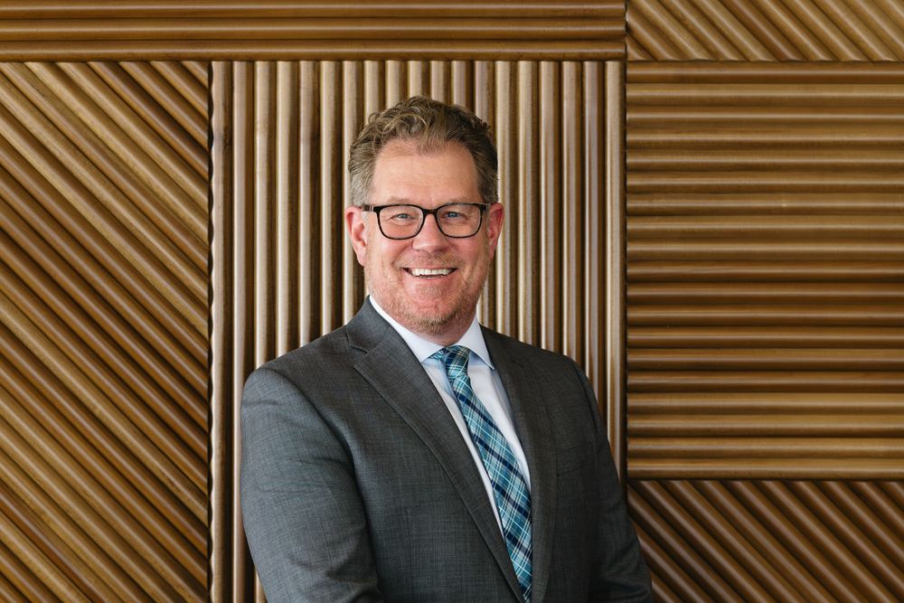 Professional wearing glasses and blue tie smiles at camera in front of wood paneled wall.
