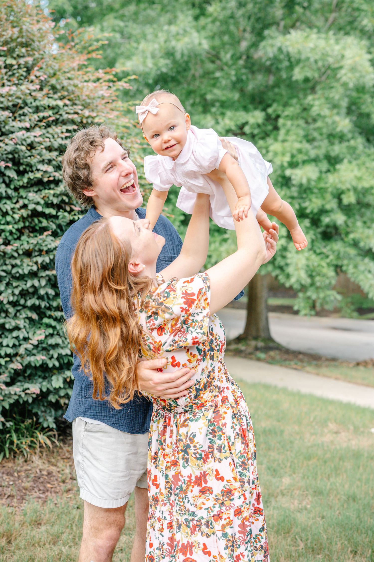 A joyful family outdoors, parents laughing as mom lifts smiling baby girl in white dress amid green trees.