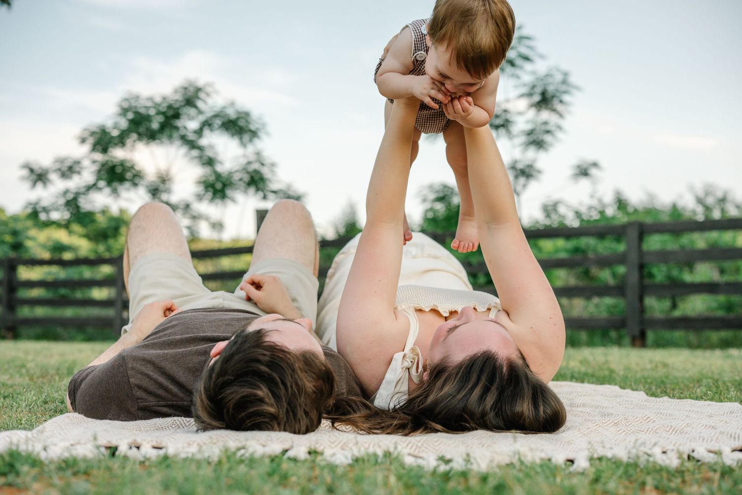 A mother lies on a blanket outdoors lifting her smiling baby in the air on a sunny day at a farm.