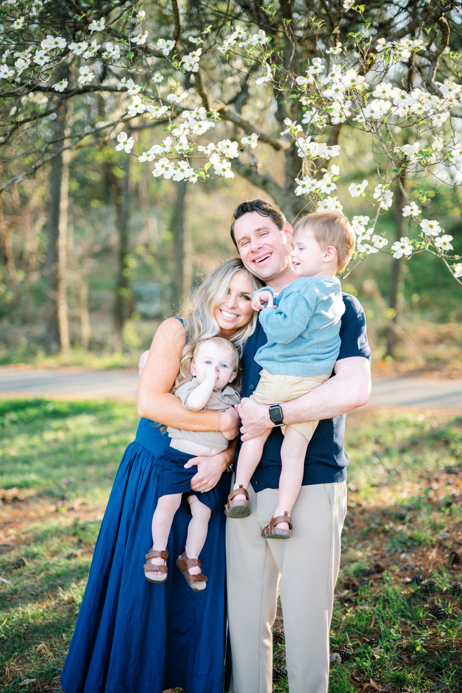 Joyful family of four posing under blooming white spring trees, parents holding two young boys.