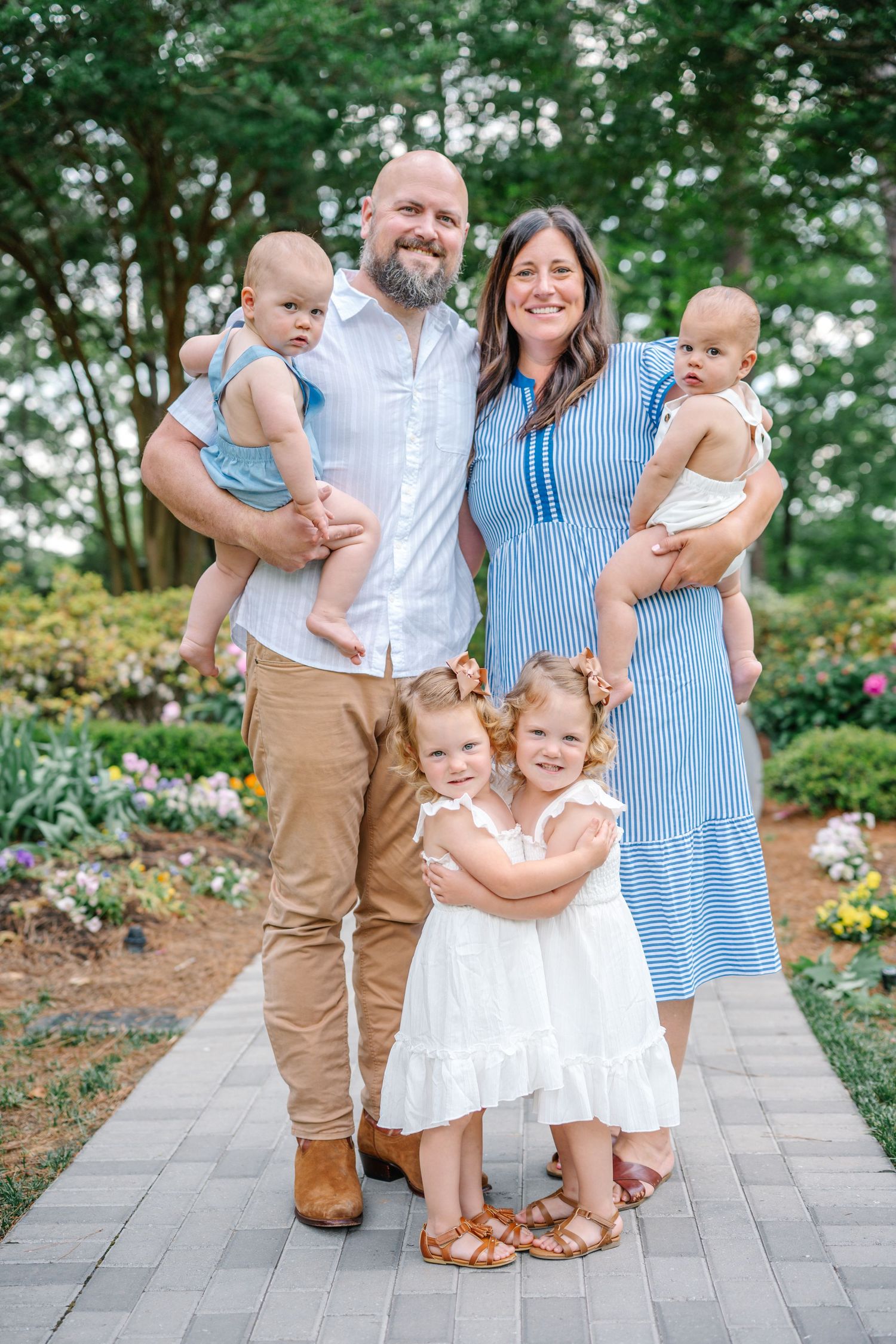 Happy family of five posing outdoors on a garden path, parents holding twin babies with two young girls in white dresses.