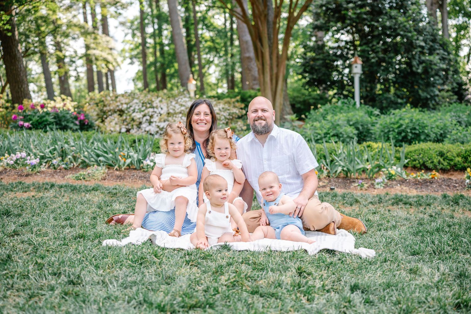 A happy family of six poses outdoors on a blanket in a lush green garden with colorful flowers in the background.