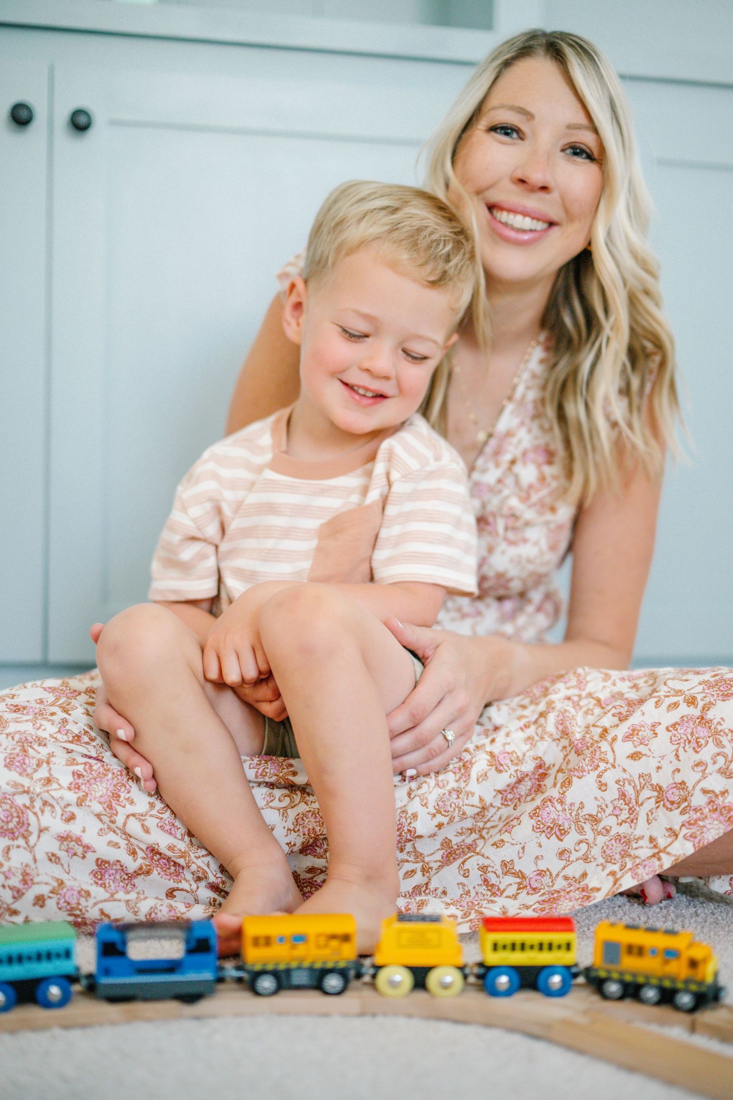 A smiling mother sits on the floor holding her young blonde toddler son, with colorful wooden toy trains in front of them.