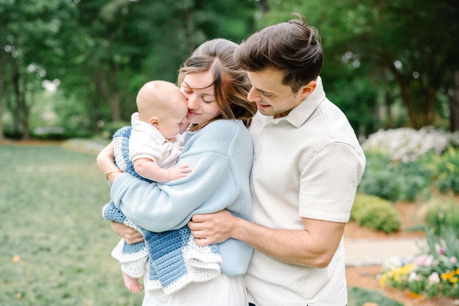 Loving parents nuzzling and embracing their baby boy in a blue knit sweater in a park.