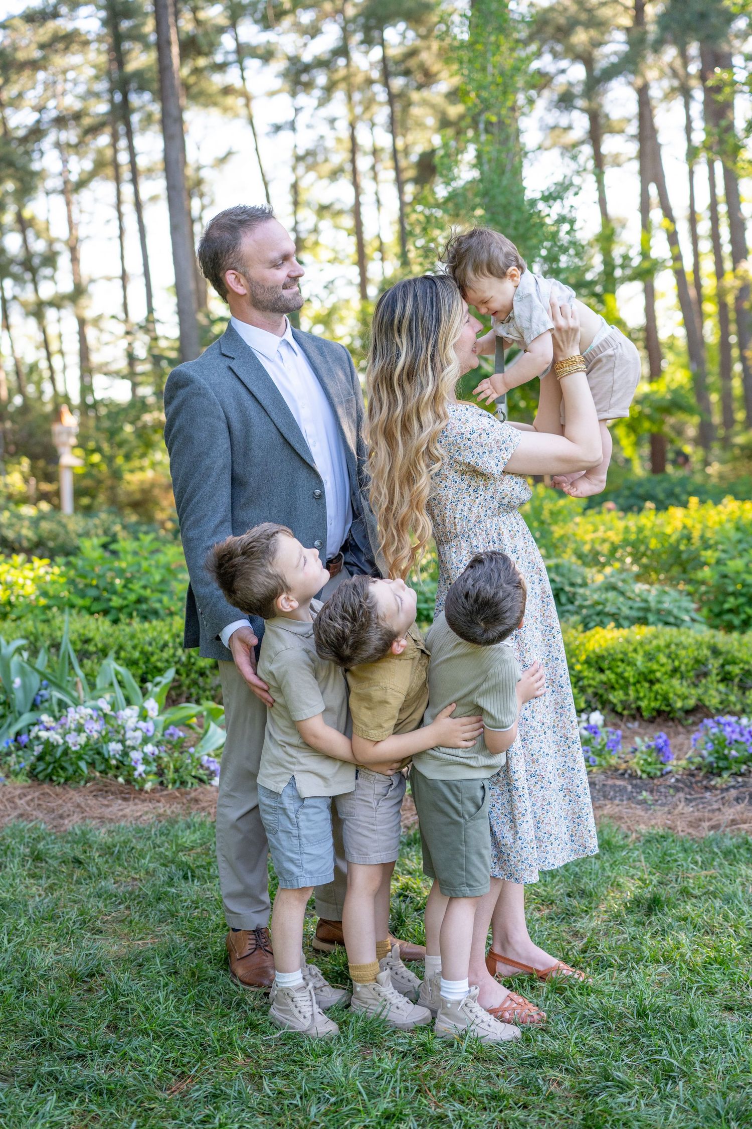 Family portrait of parents with four young children hugging in a lush garden with tall pine trees and colorful flowers.