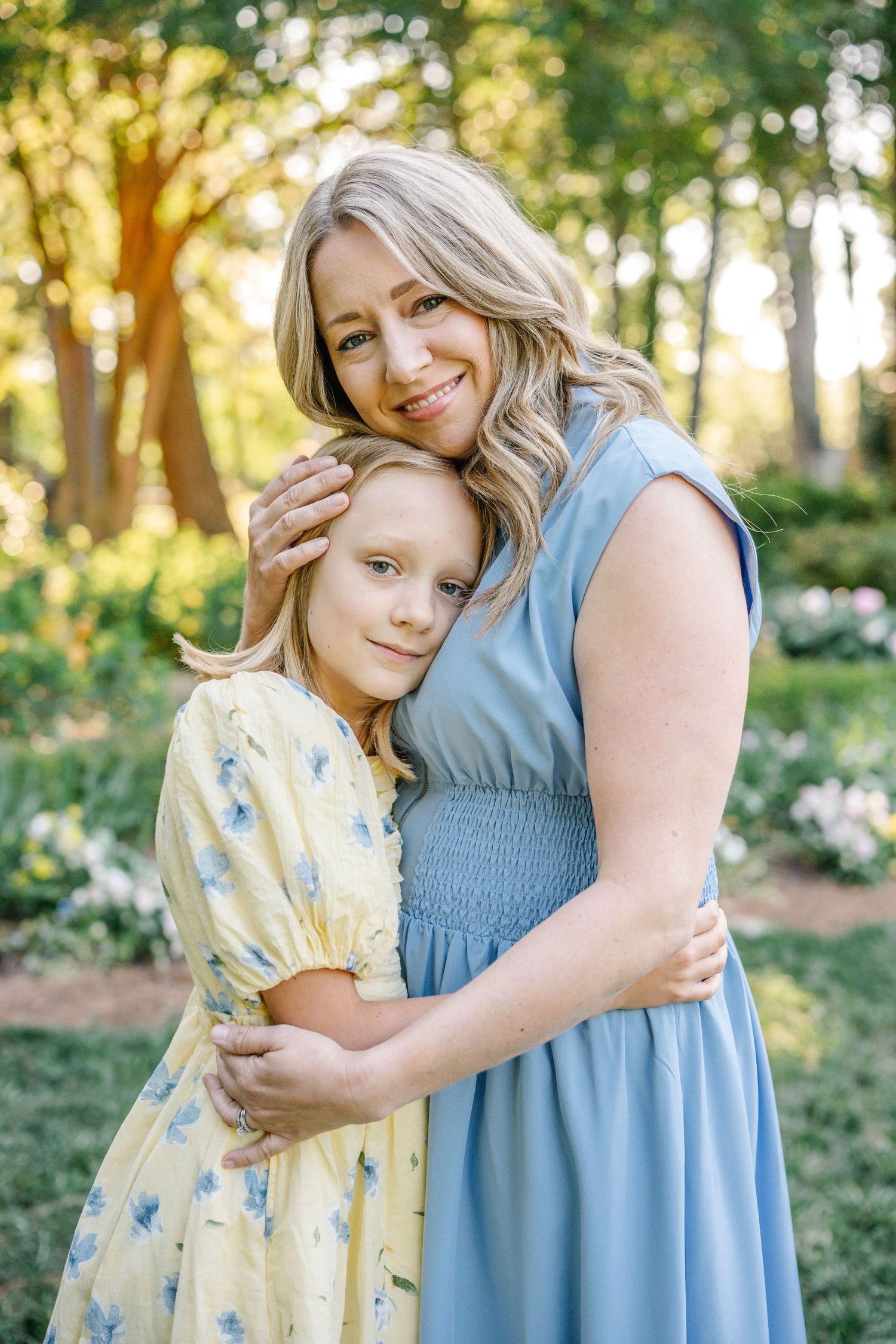 A woman in a blue dress hugs a young girl in a yellow floral dress outdoors in a sunlit garden.
