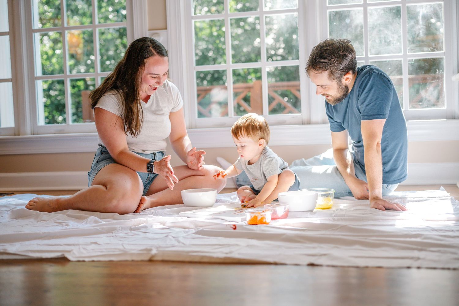 Family sitting on a blanket on a hardwood floor, sharing a meal together in a bright, sunlit room with large windows.