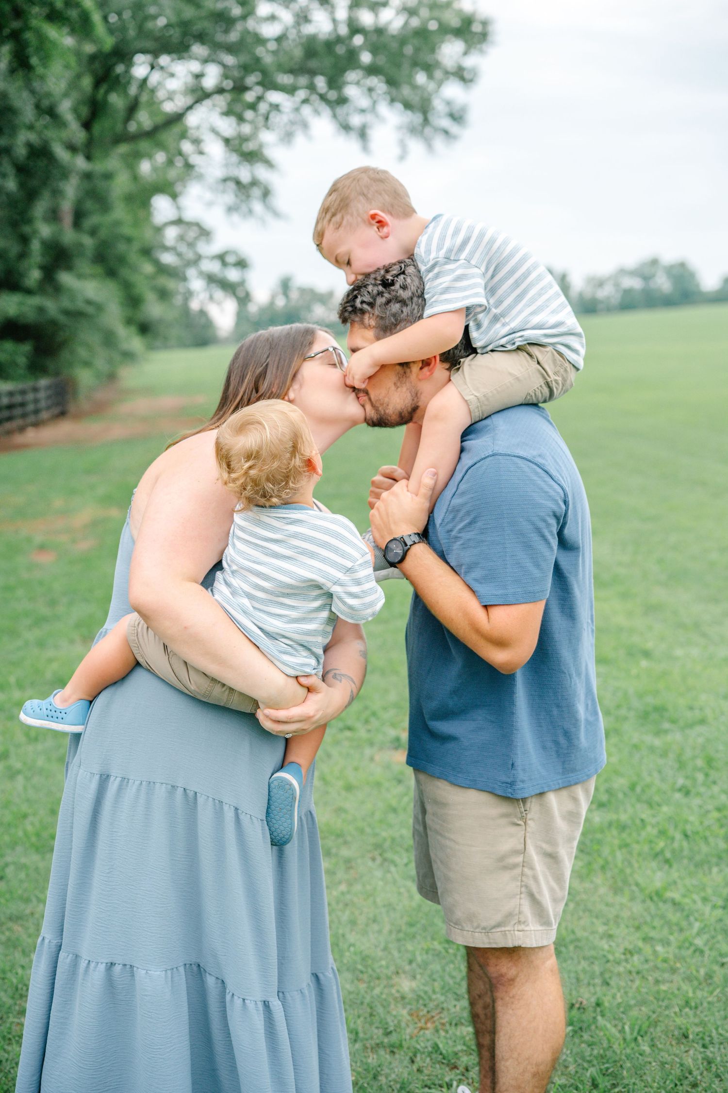 A family of four shares a sweet kiss outdoors, with mom holding a baby and dad carrying a toddler on his shoulders.