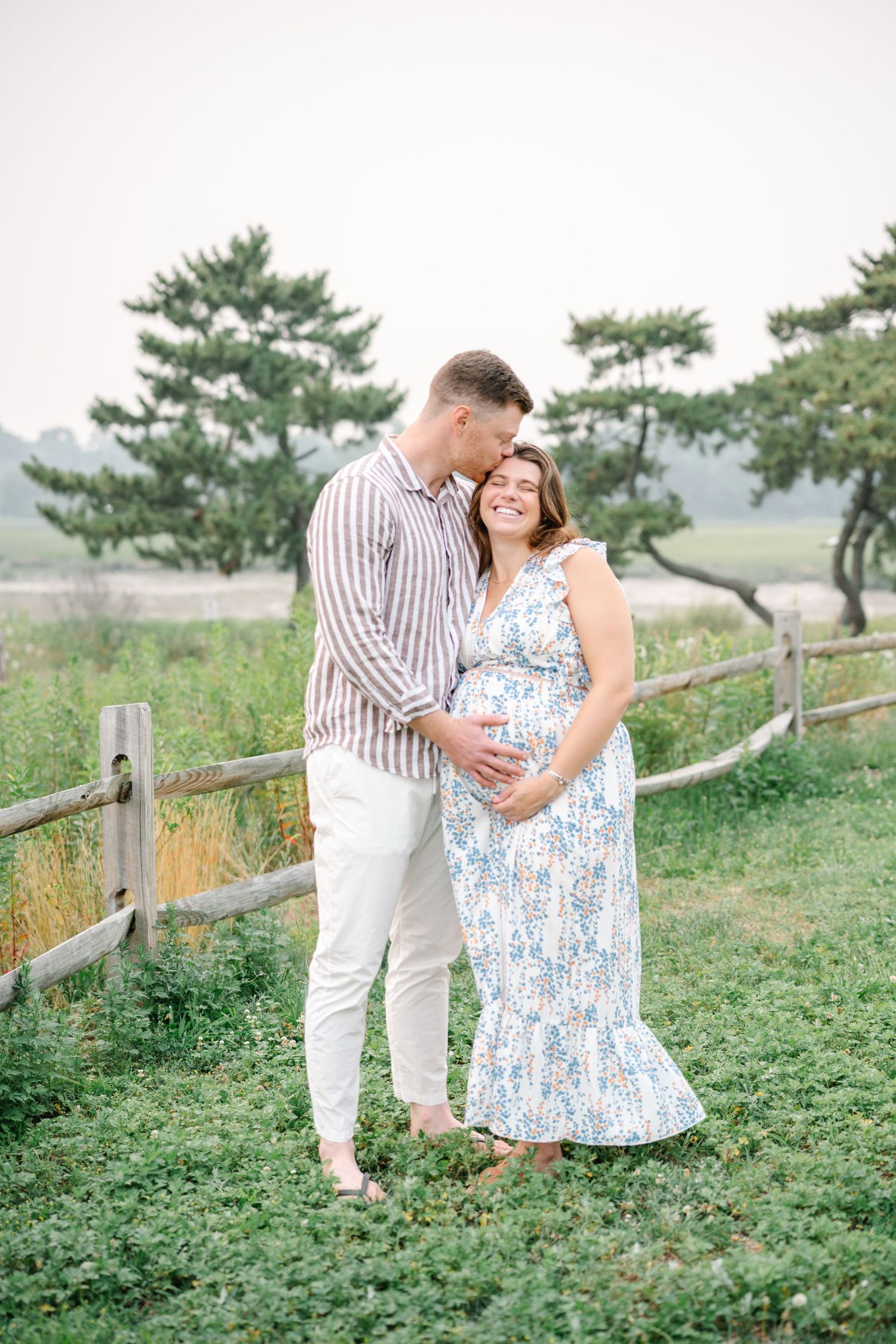 Couple poses outdoors by rustic fence, woman in floral maternity dress, man in striped shirt, both cradling baby bump.