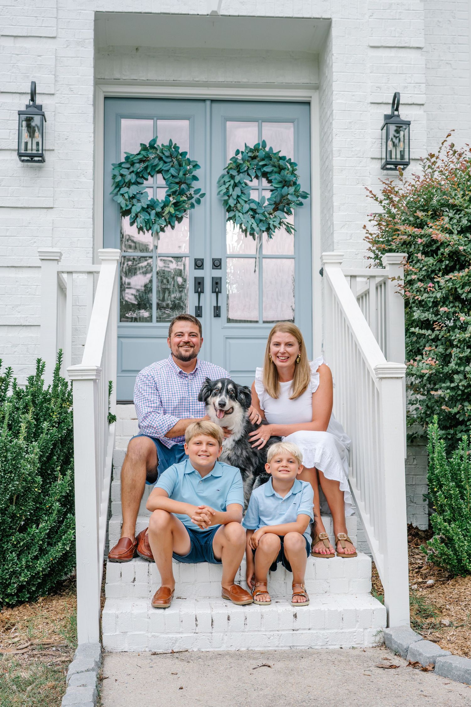 Family of four with dog posing on white brick home steps with blue double doors and green wreaths.
