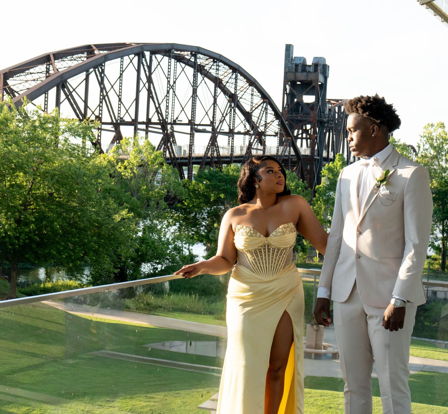 A couple in formal attire poses outdoors near a bridge, wearing a gold gown and white suit with boutonniere.