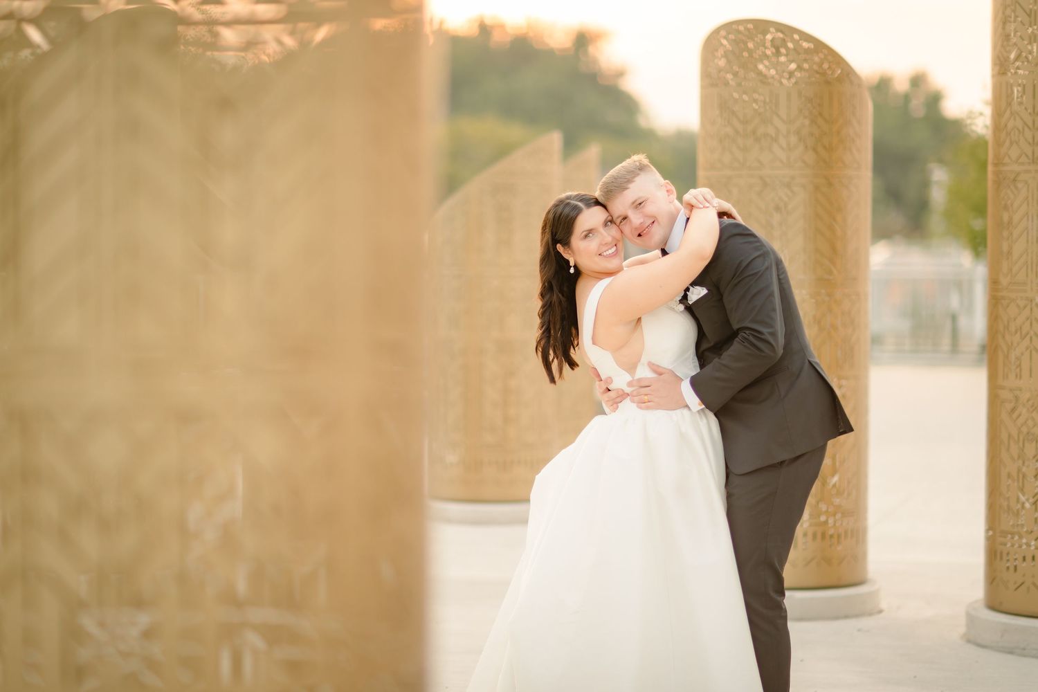A romantic wedding couple embraces at sunset near elegant stone columns in an outdoor setting.