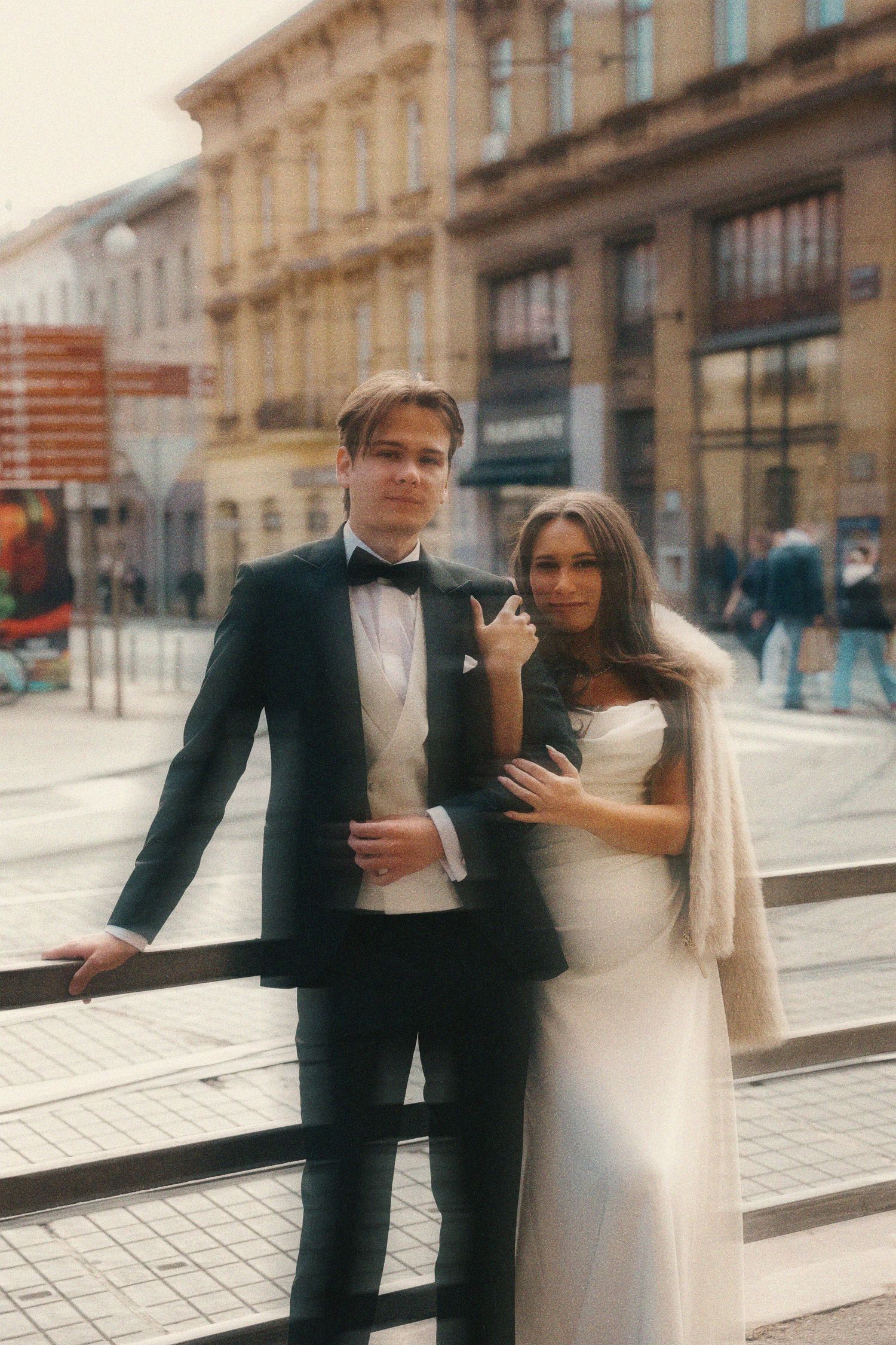 A romantic couple in formal attire embraces on a European city street with elegant architecture in the background.