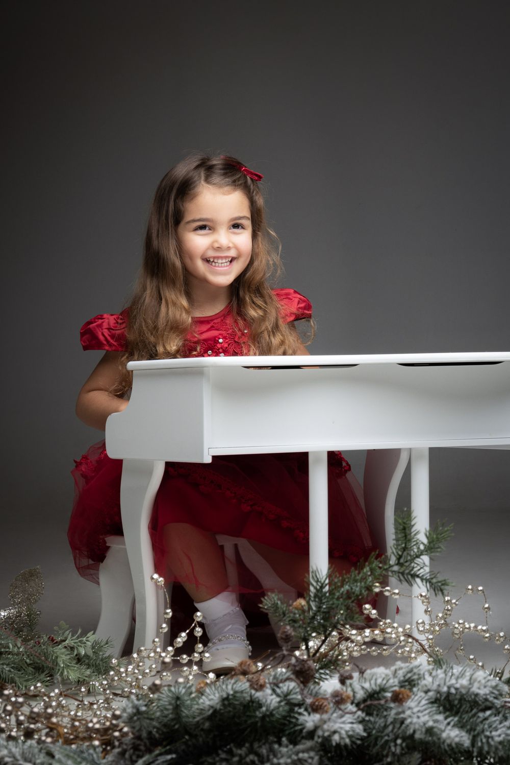 A child in a red dress sits at a white piano surrounded by winter greenery and baby's breath flowers.