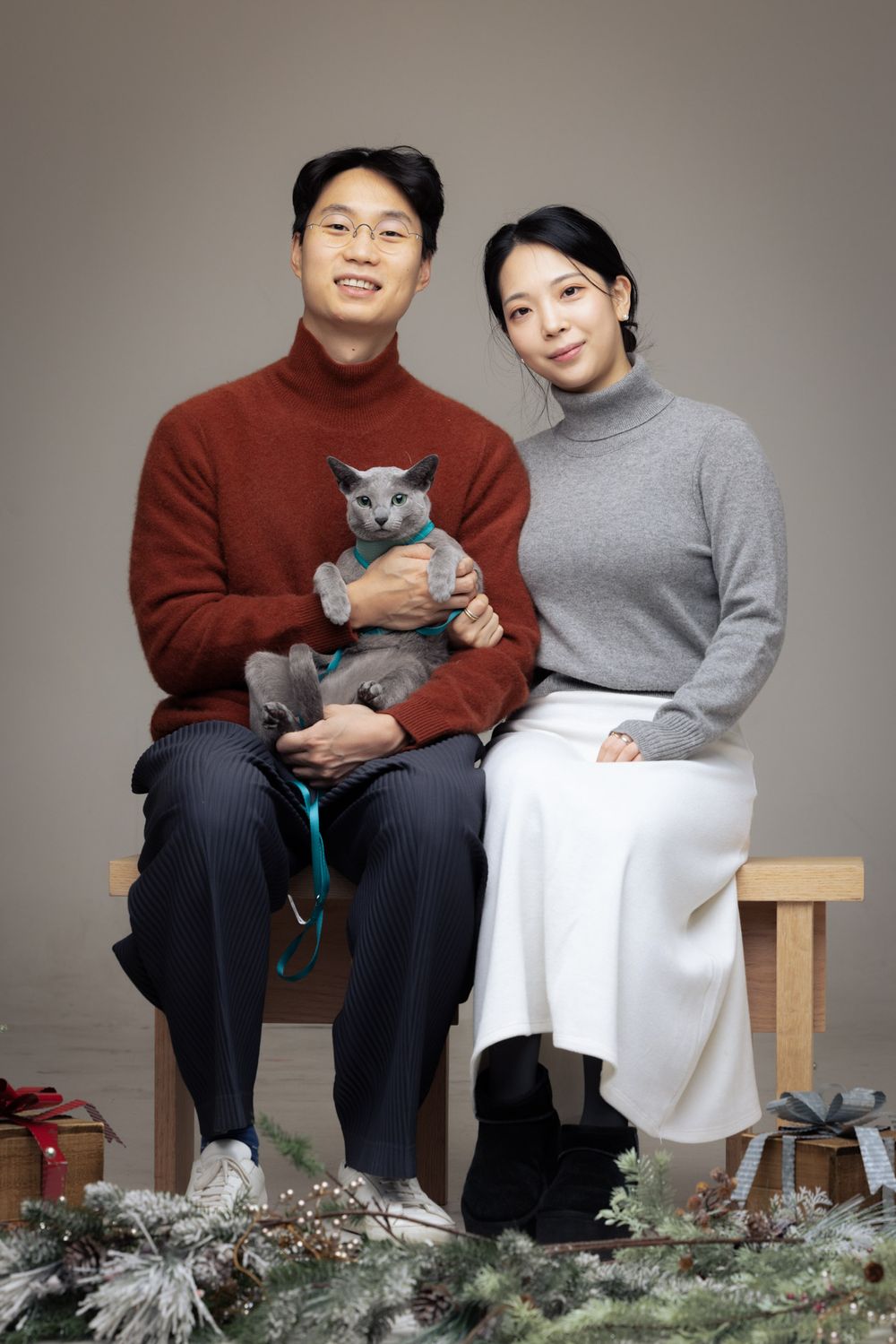 A couple sits on a wooden bench with their gray cat while posing for a series of holiday photos with greenery decorations.
