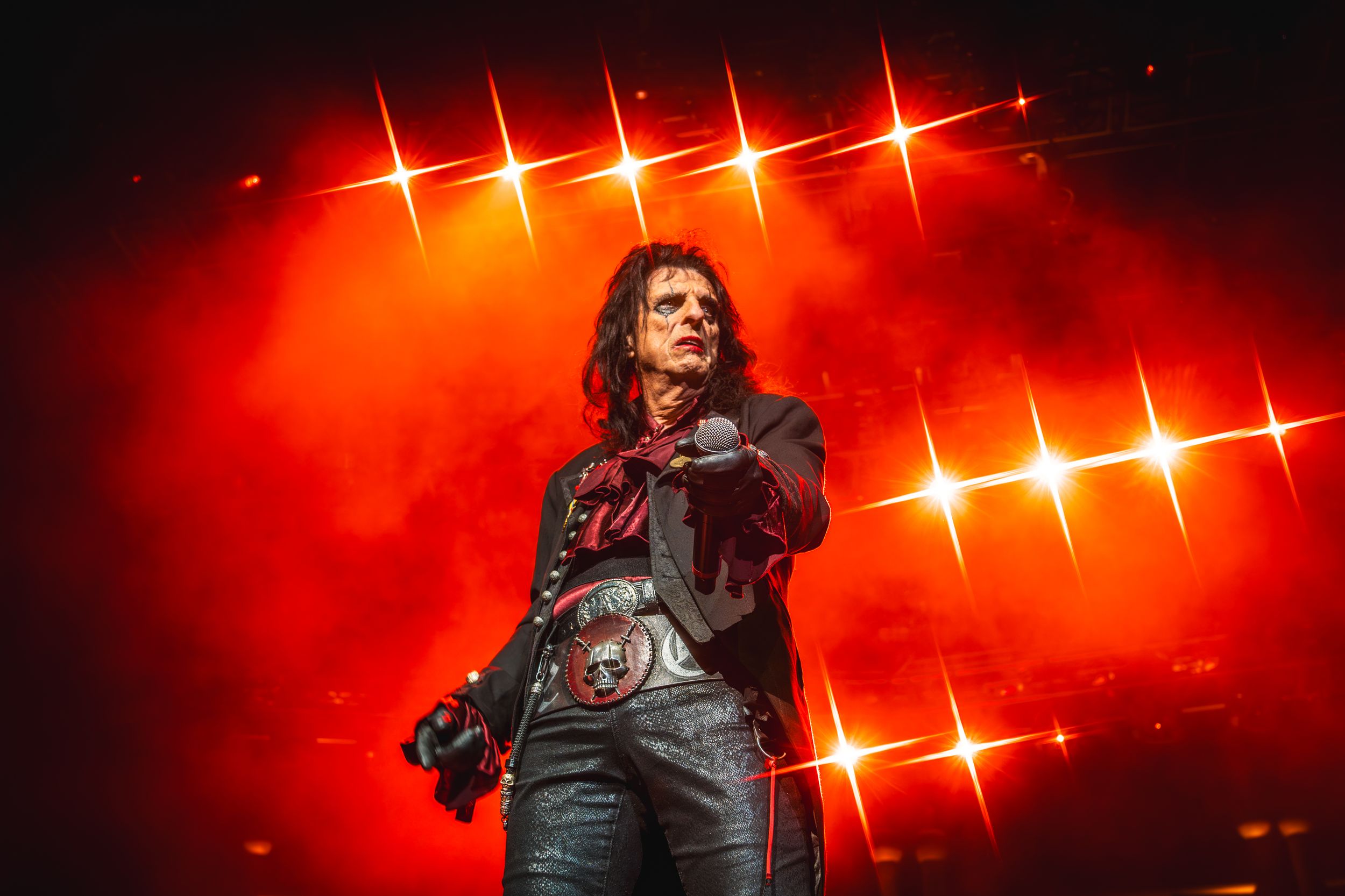 Rock performer in leather jacket sings passionately on stage with dramatic red lighting and star-like effects in background.