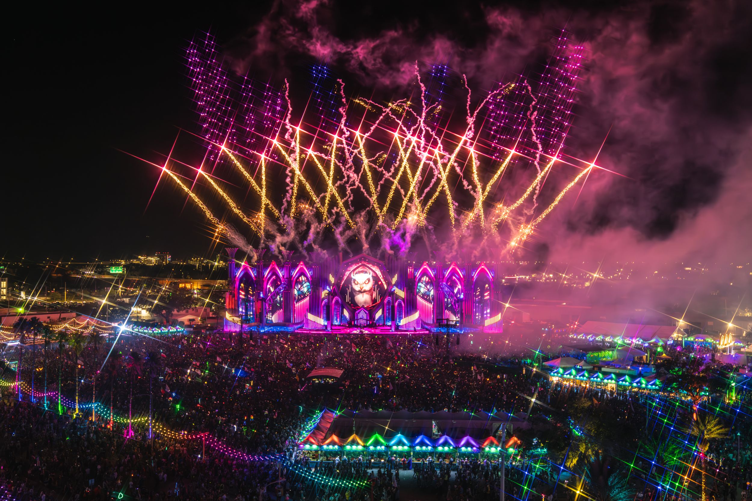 Colorful fireworks display illuminates a festival stage with vibrant purple and pink lights against the night sky.