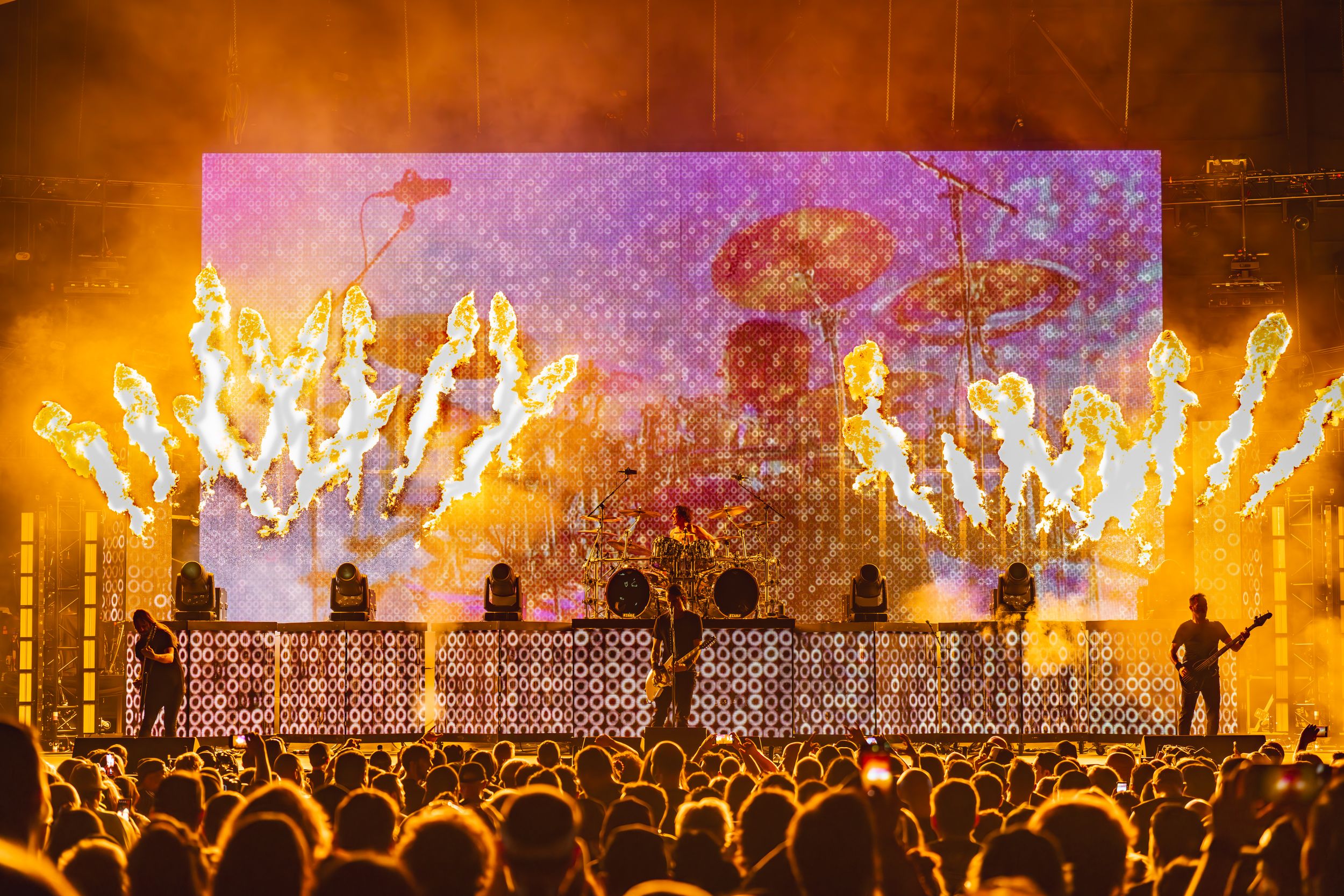 Concert stage illuminated with bright orange flames and pyrotechnics as silhouetted crowd watches the performance.