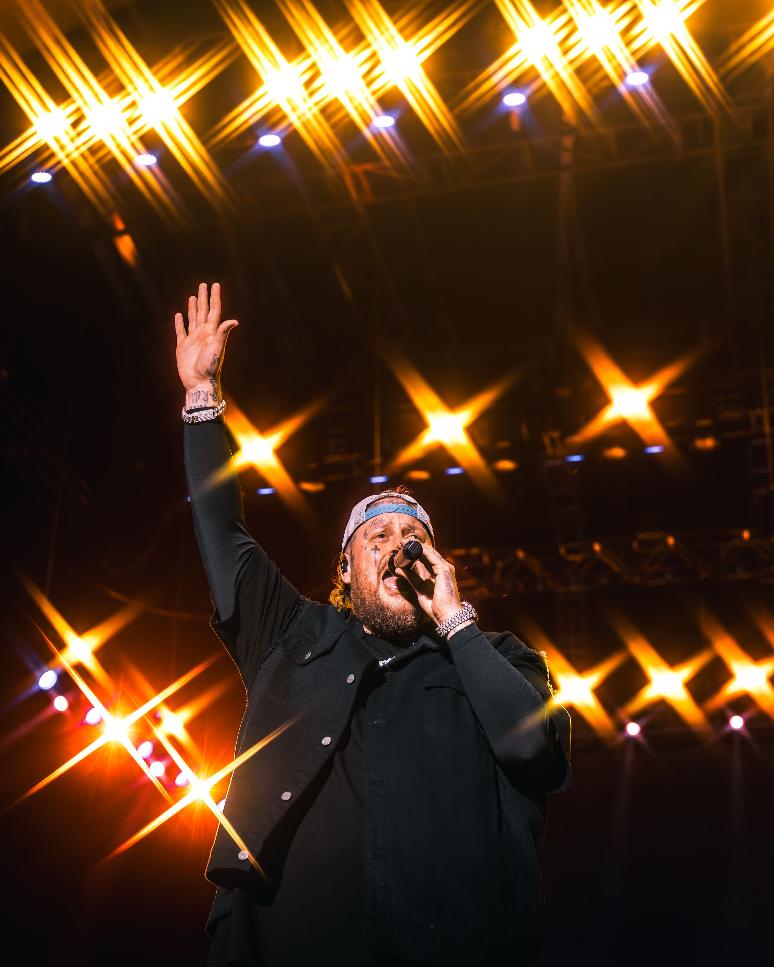 A performer in black clothing raises their hand while singing on stage with dramatic gold star-shaped lighting effects in background.