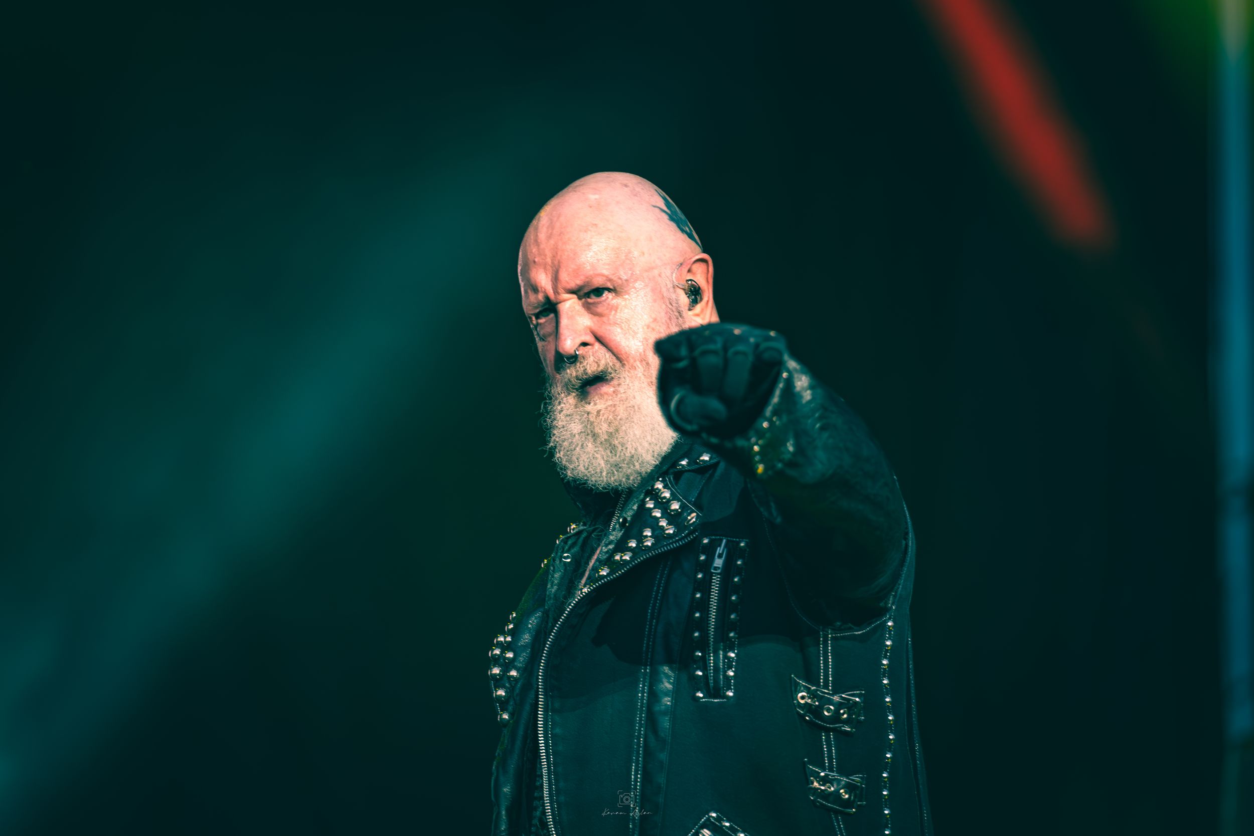 Heavy metal performer in black leather jacket singing on stage with dramatic teal lighting in the background.