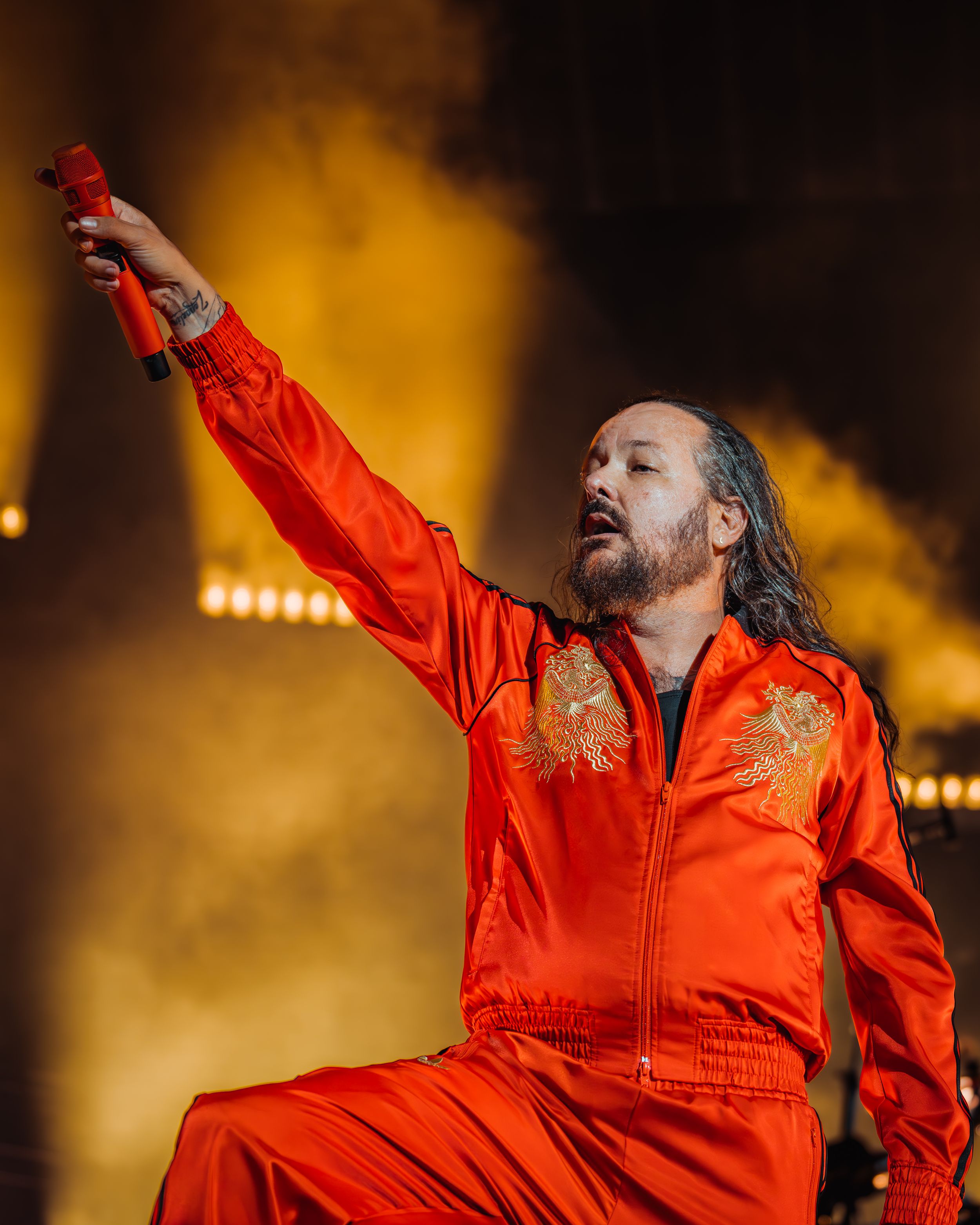Performer in bright orange jumpsuit strikes dramatic pose against dark smoky background during stage performance.