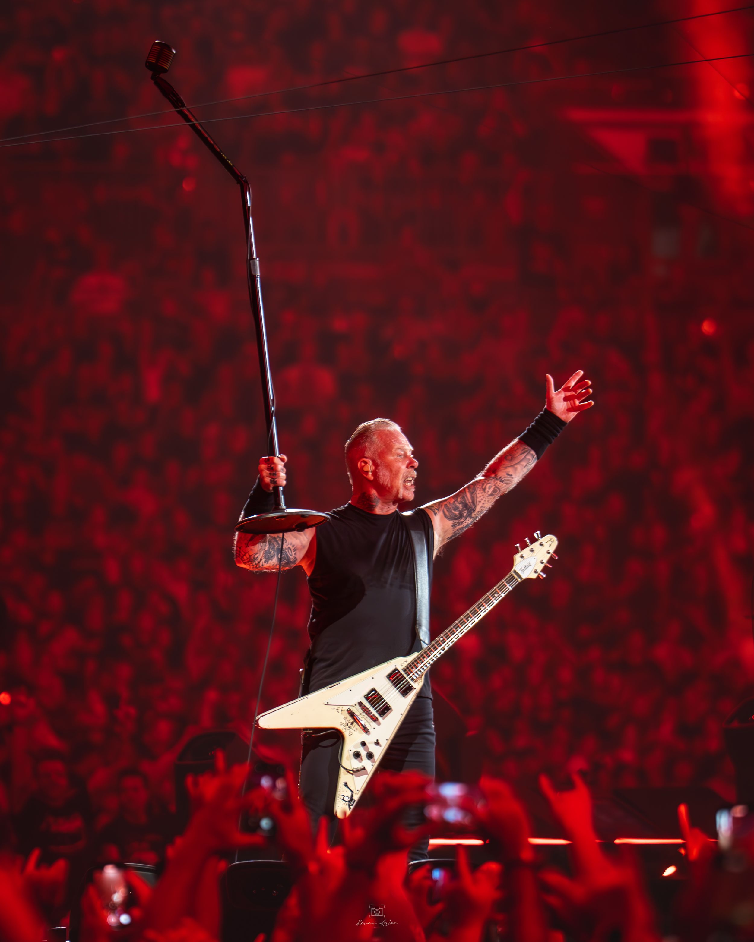 A performer plays guitar on stage with dramatic red lighting and silhouetted crowd in the background.