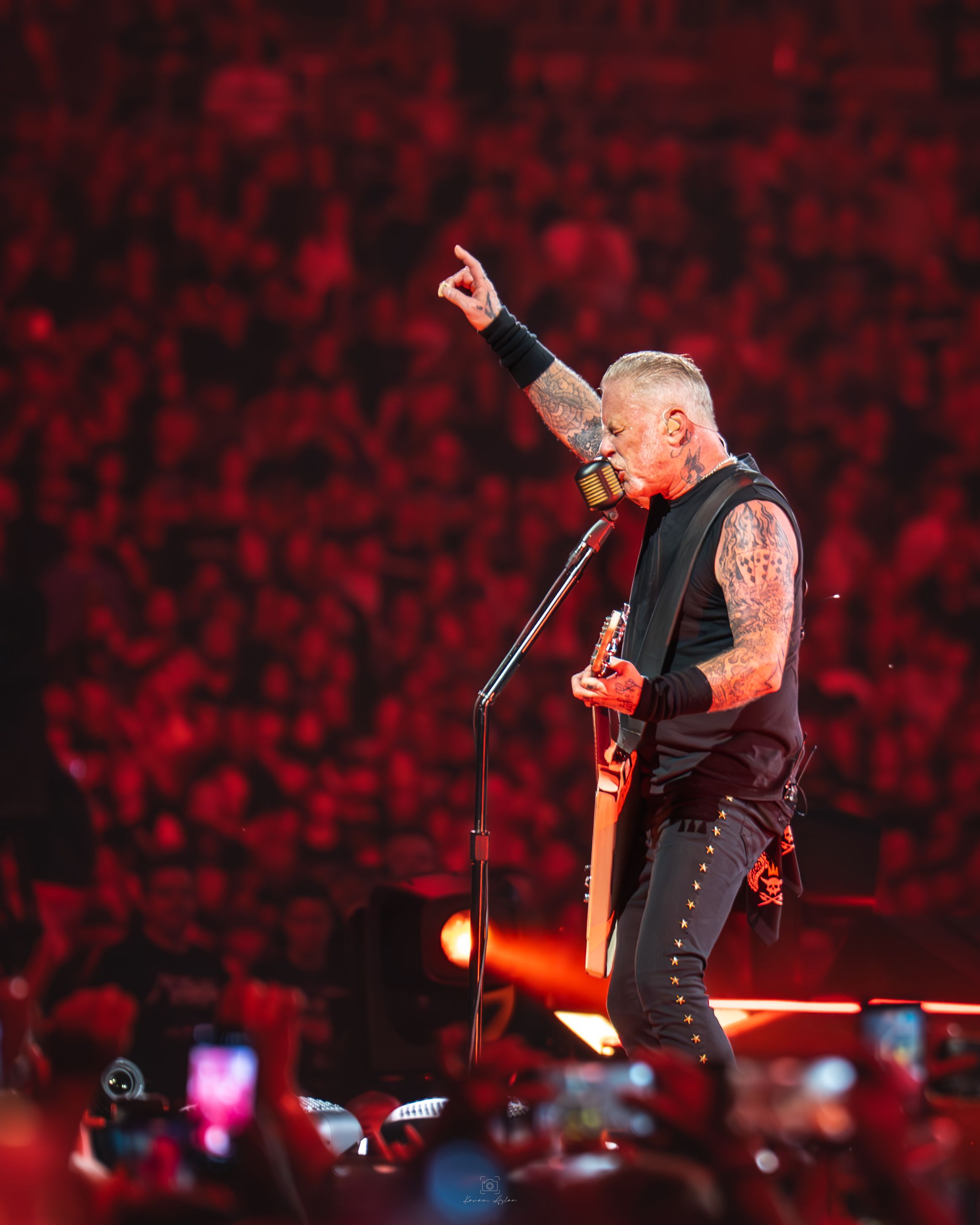 Heavy metal musician performs on stage with guitar against dramatic red lighting and crowd holding phones.