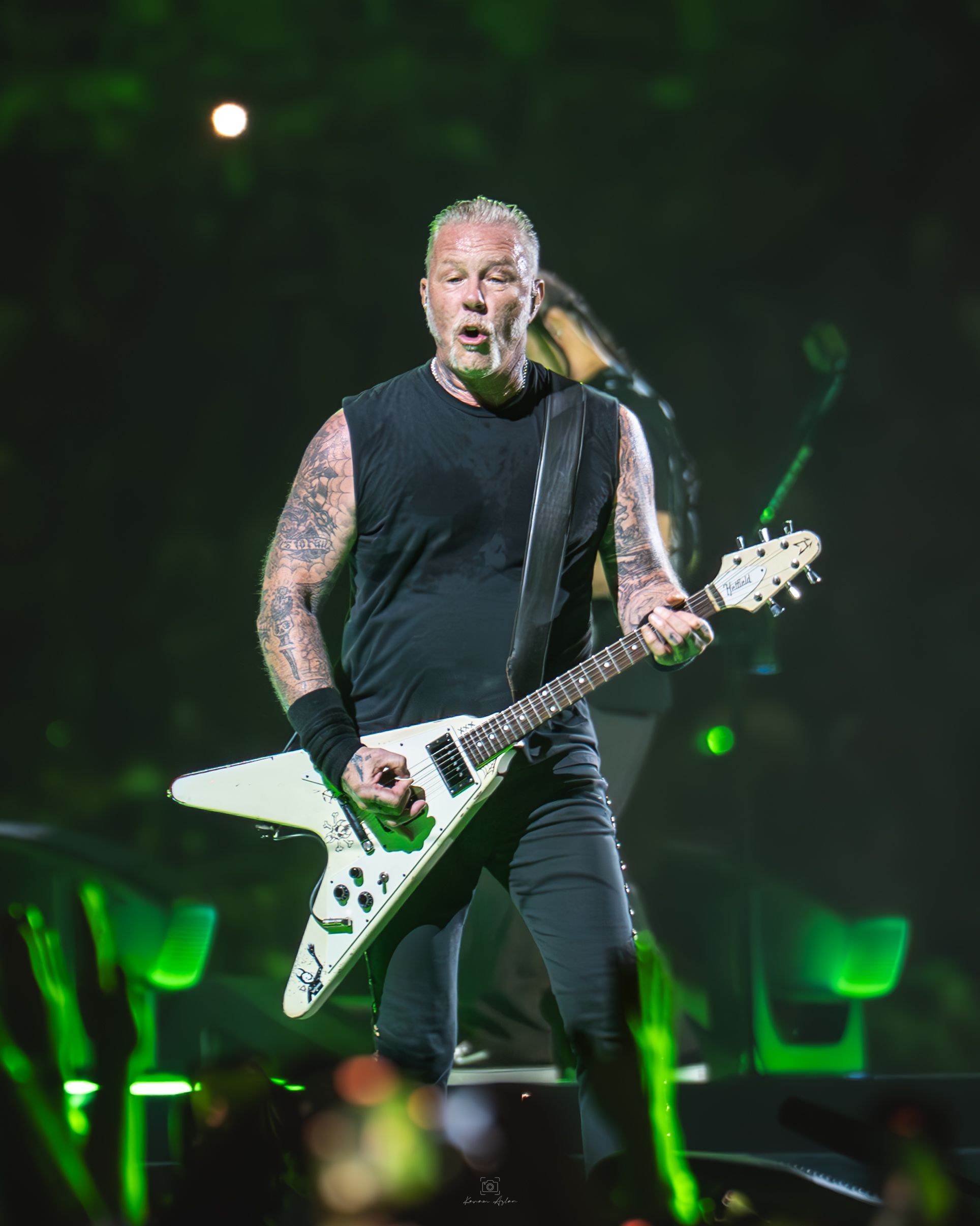 A musician performs on stage with a white Flying V guitar under green stage lights and a dark backdrop.