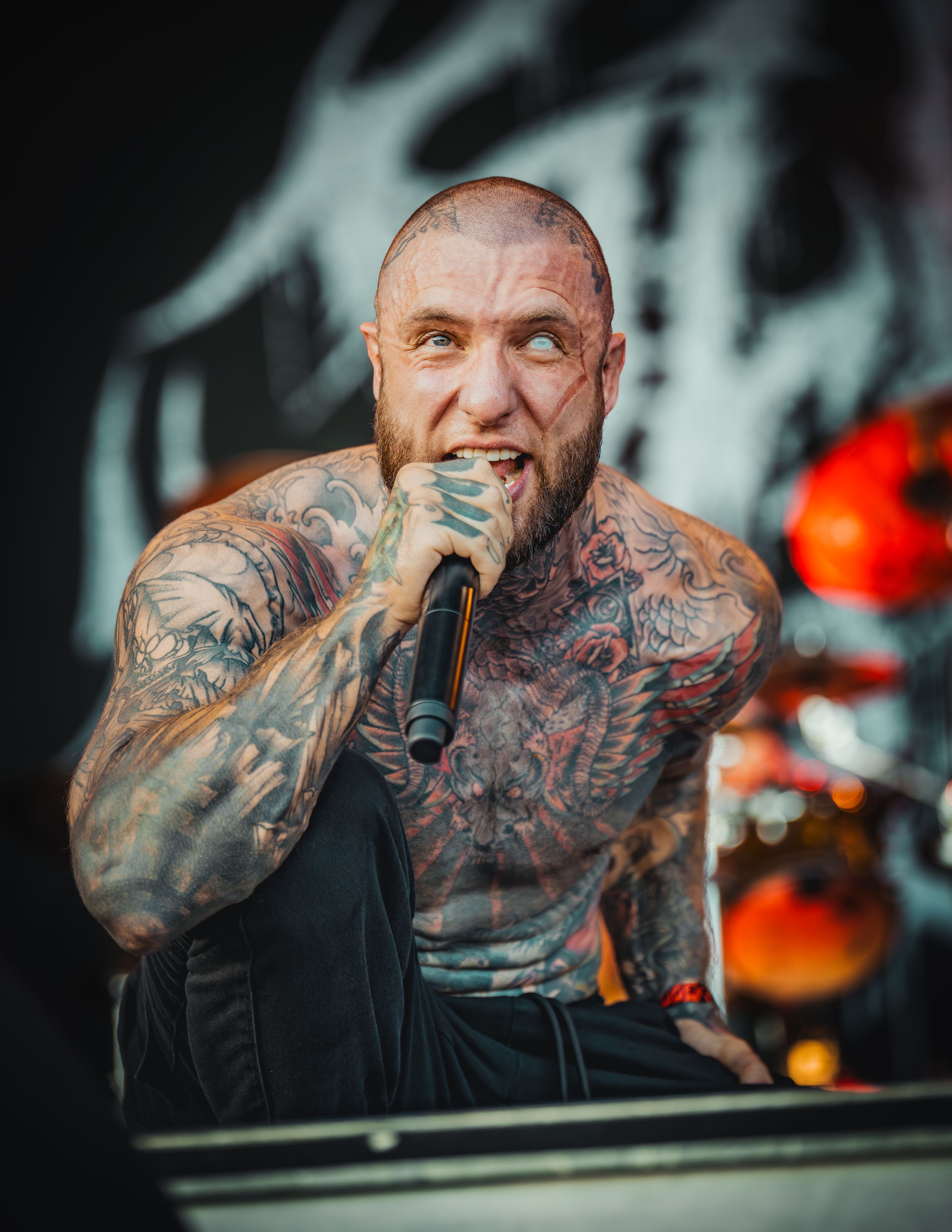 Heavily tattooed performer singing passionately on stage with dramatic lighting and dark background.