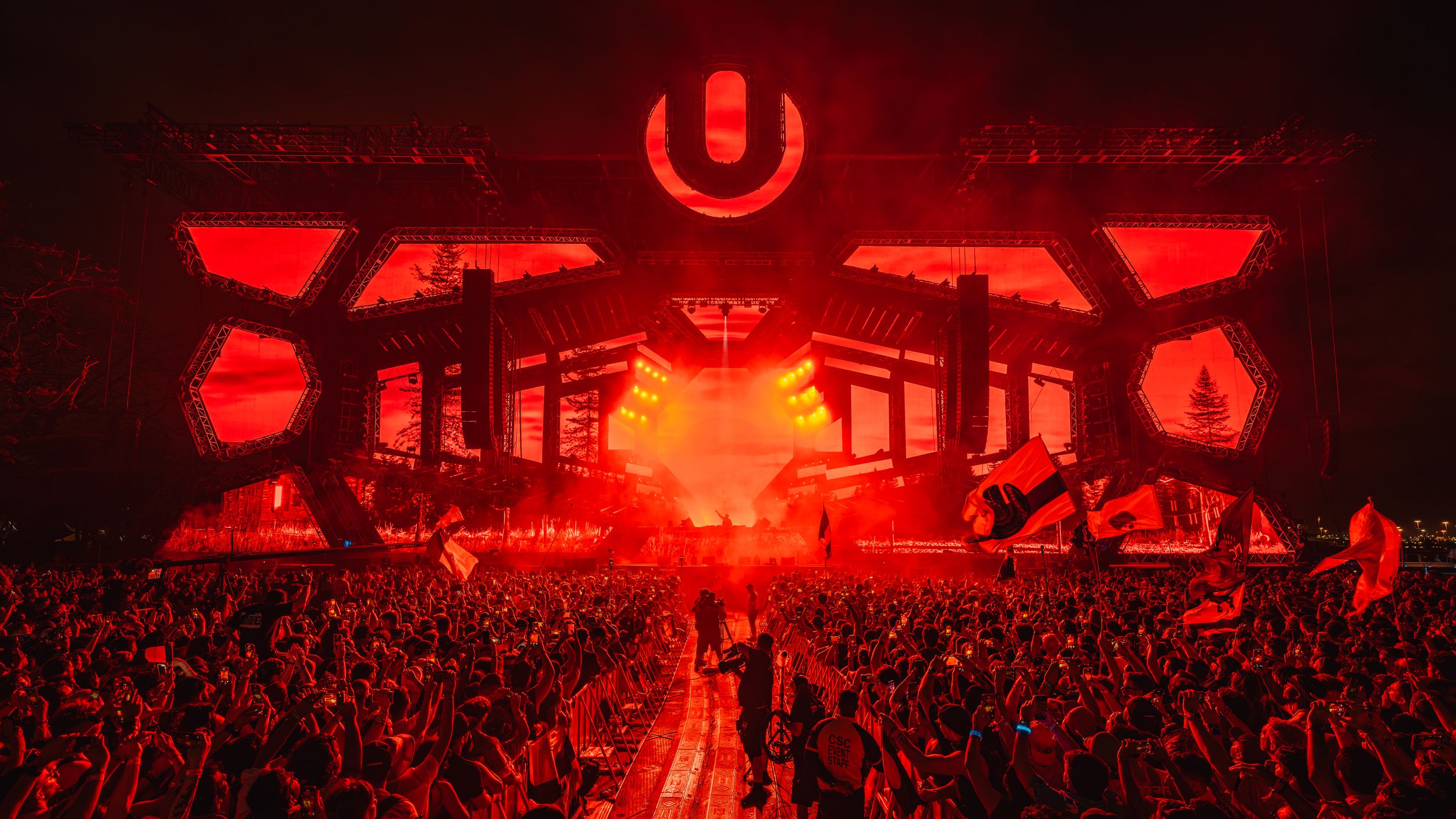 Concert crowd illuminated in dramatic red lighting at a large arena venue during a live performance.