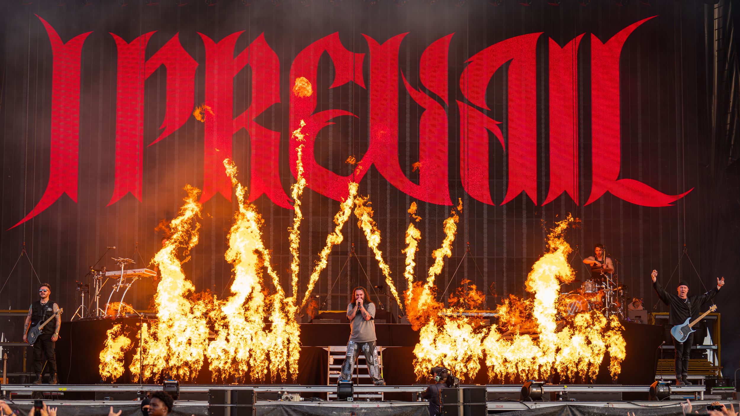 Heavy metal band performs on stage with dramatic fire effects and red illuminated logo backdrop during intense concert.