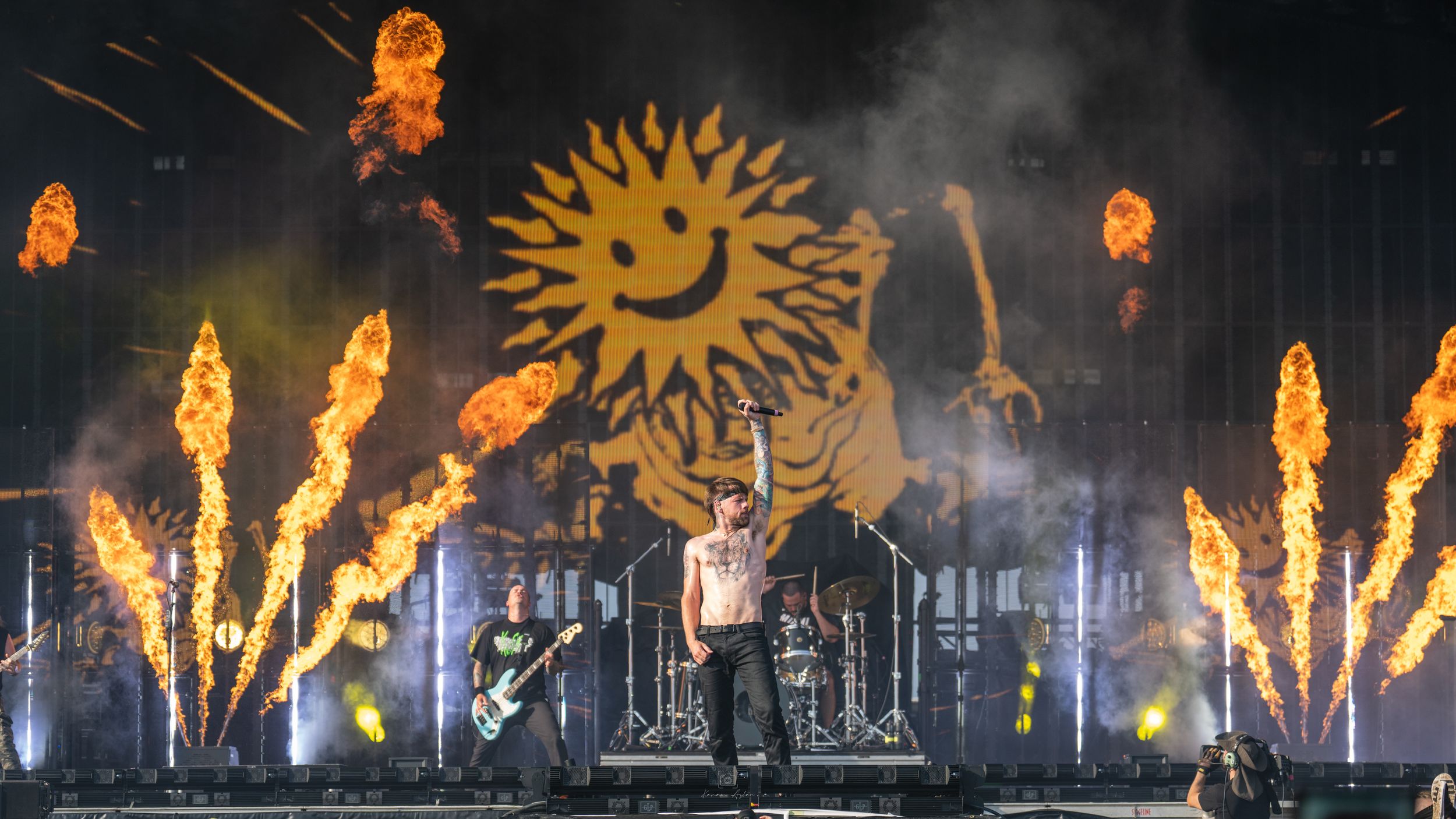 Illuminated stage performance with fire effects and a large sun-shaped backdrop glowing against a dark nighttime setting.