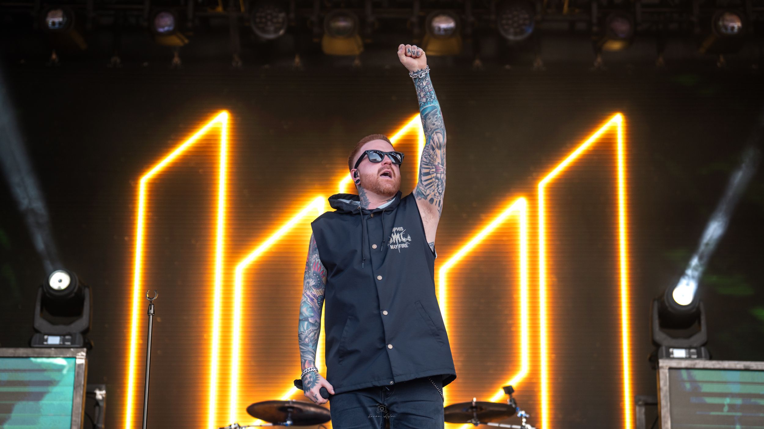 A performer in a black outfit stands on stage with bright yellow neon light bars creating geometric patterns behind them.