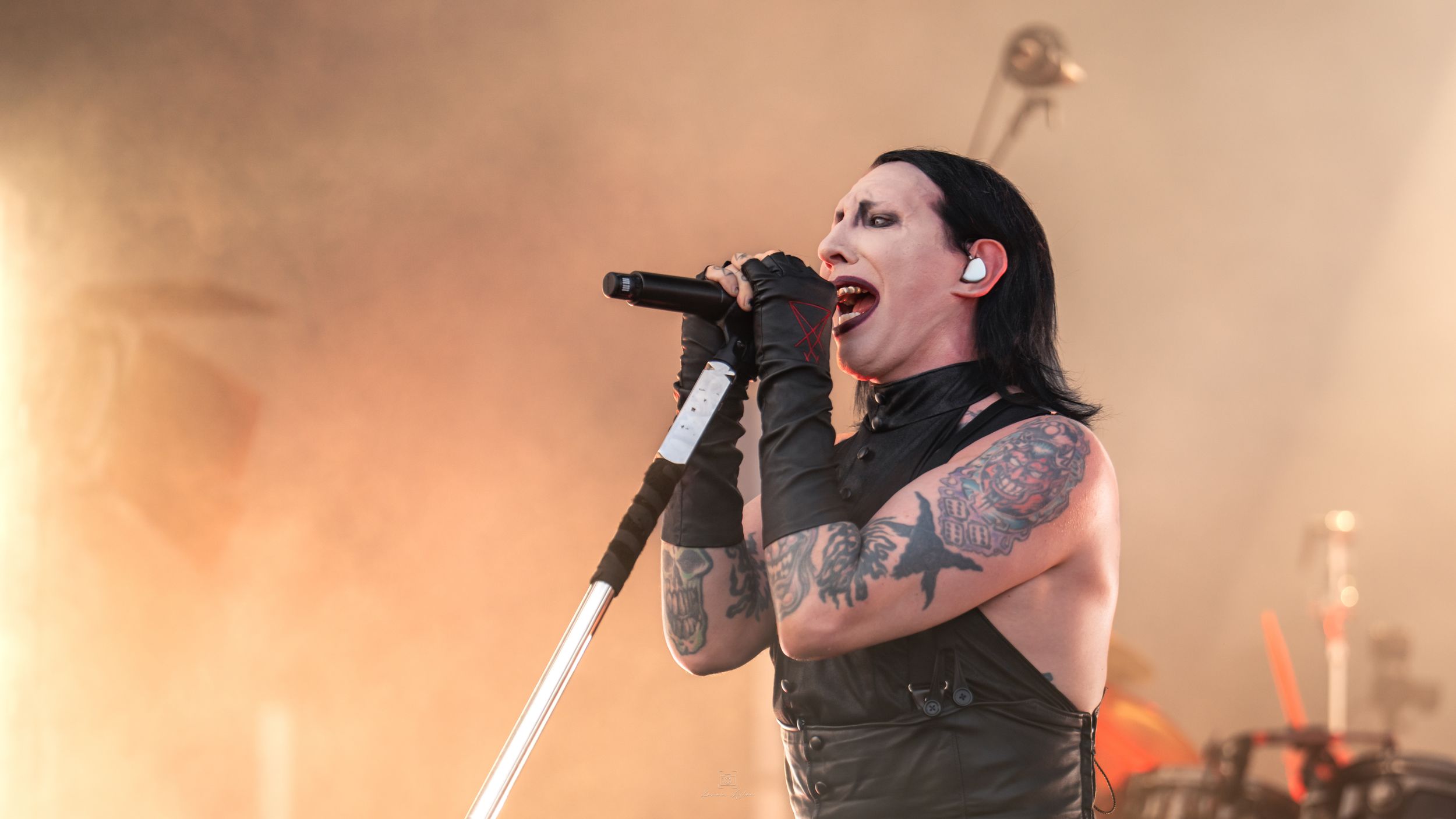 A performer in a dark vest sings passionately into a microphone on stage with dramatic orange lighting behind them.