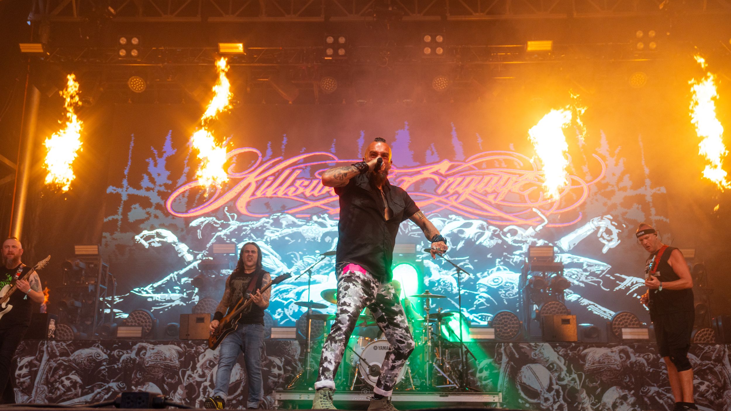 Concert stage with bright flames shooting upward and colorful neon lighting effects creating a dramatic atmosphere.