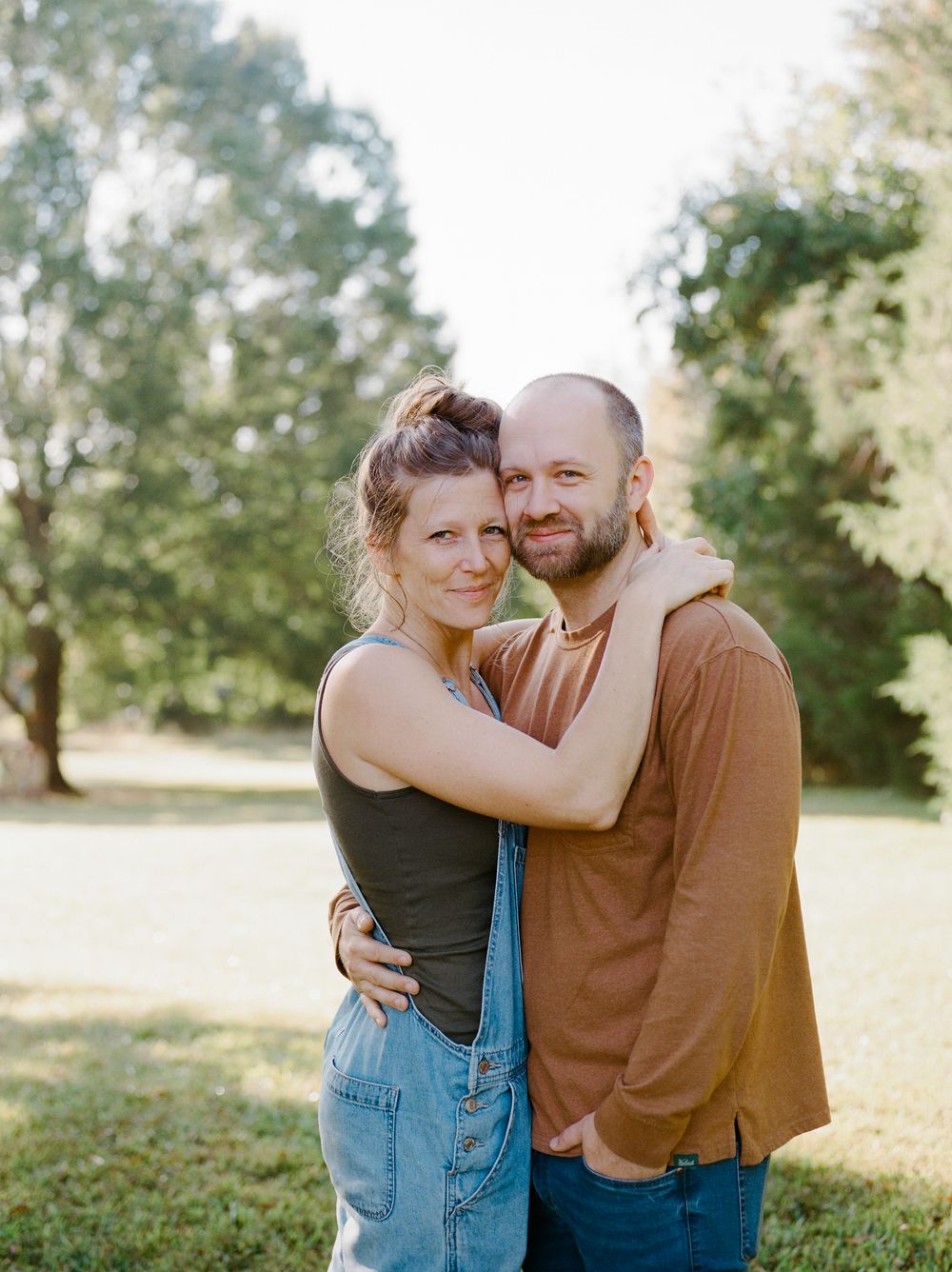 A couple embraces outdoors on a sunny day in a park with trees in the background.