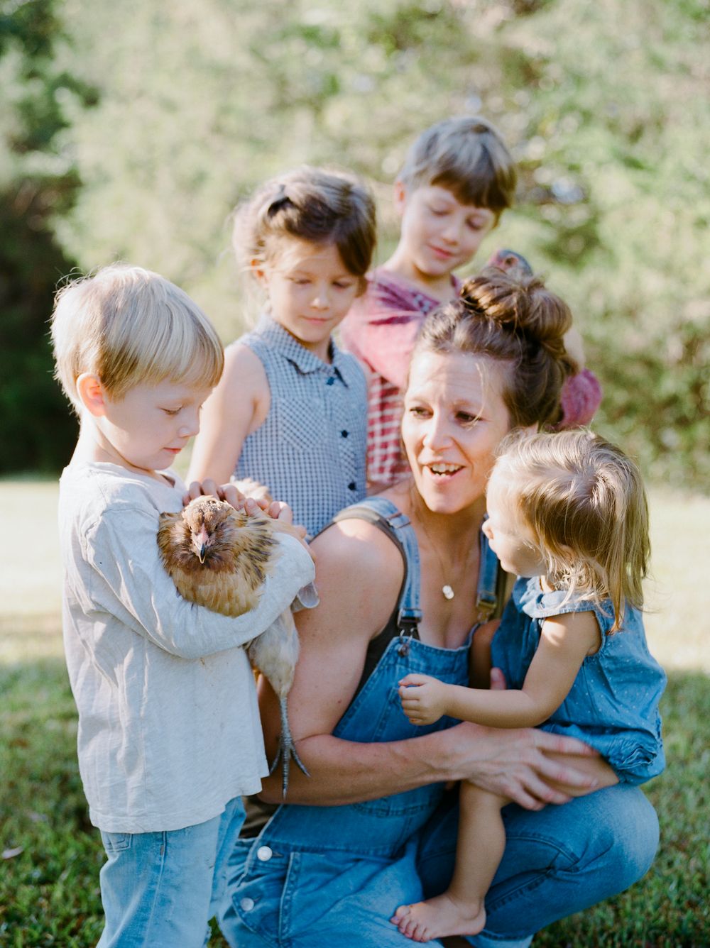 A group of children and an adult in overalls gather together outdoors on a sunny day.