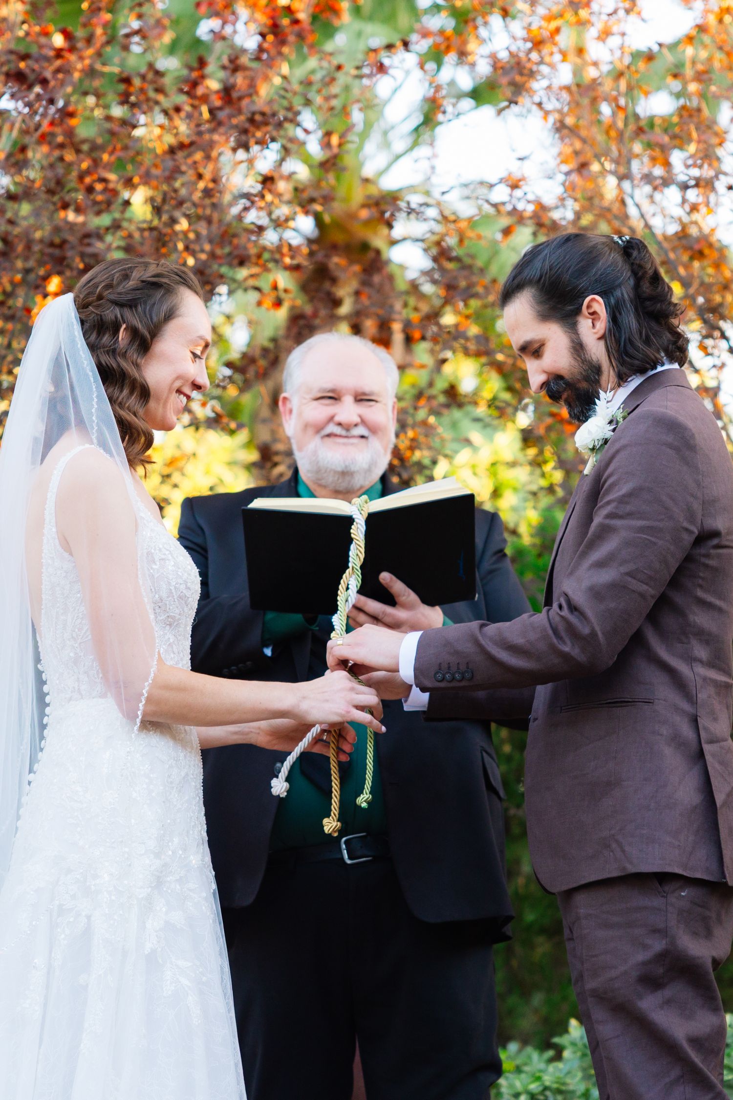 A wedding ceremony takes place outdoors in autumn with colorful fall foliage in the background.