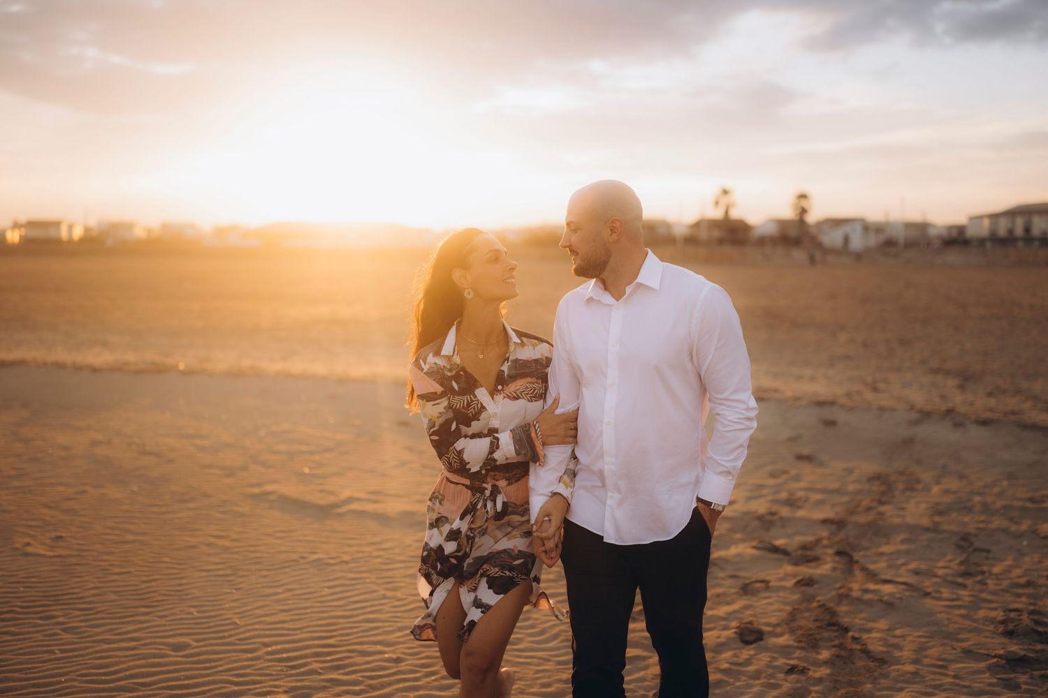A couple walks hand in hand along a sandy beach at sunset with golden light illuminating their silhouettes.