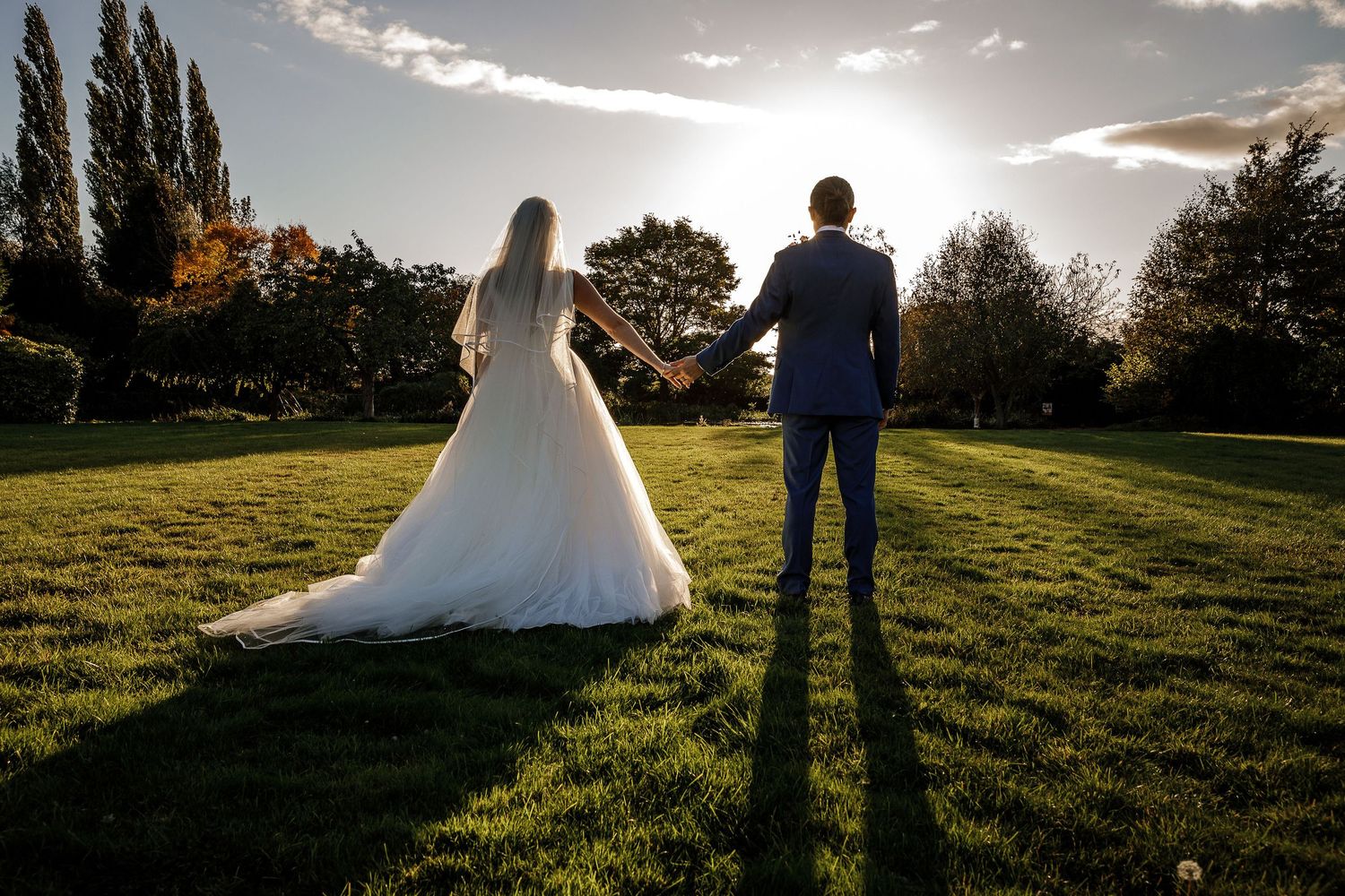 Bride and groom holding hands in a romantic outdoor setting with soft sunlit trees and foliage at Hodsock Priory