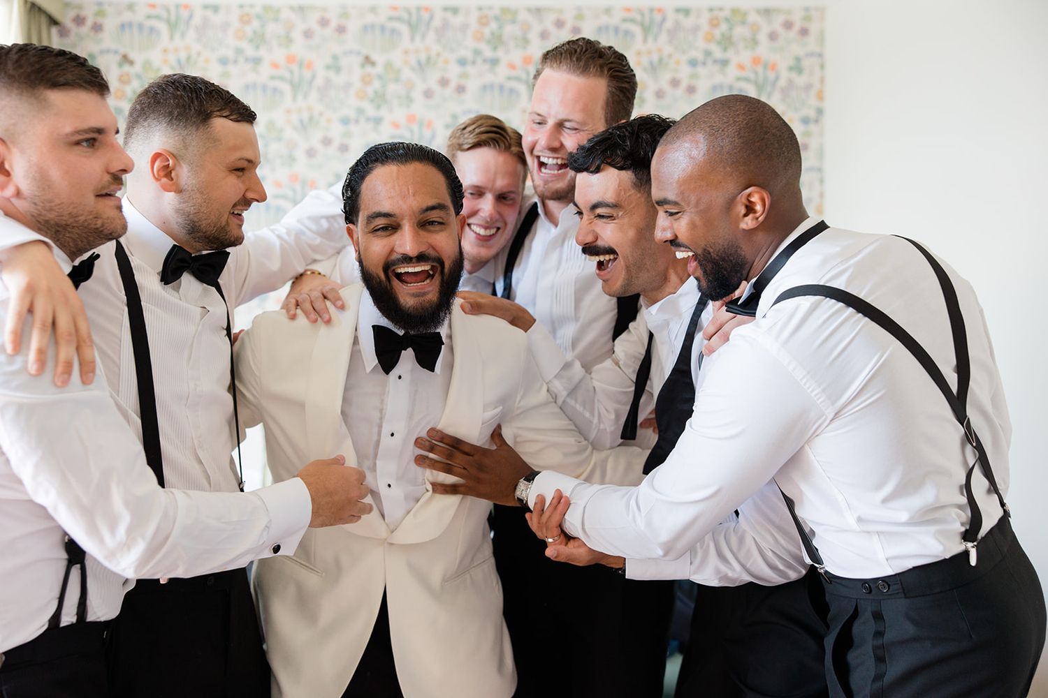Groomsmen in white shirts and suspenders share joyful laughter during wedding celebration.