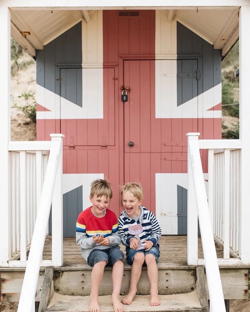 Two young children sitting on the steps of a colourful union jack painted beach hut in summer.