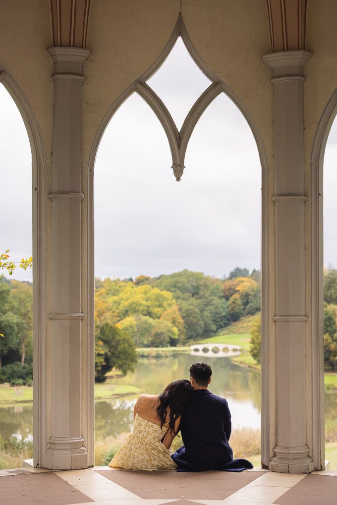 A couple sits together in a romantic stone gazebo overlooking the serene lake and autumn landscape of Painshill Park in Surrey.