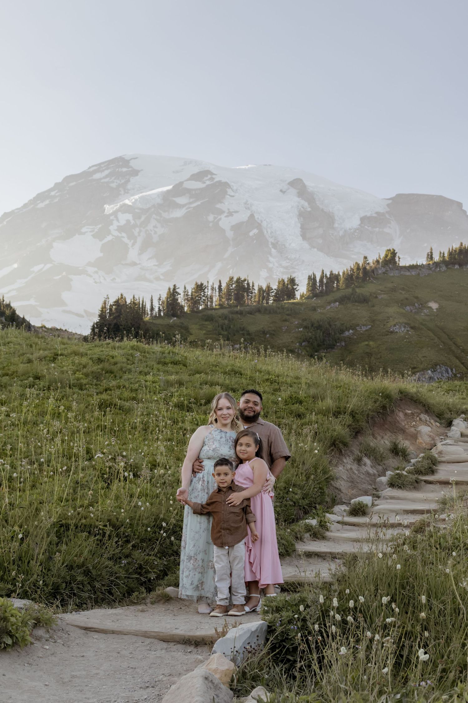 Family of four posing on a mountain trail with snow-capped Mount Rainier and green meadows in the background.